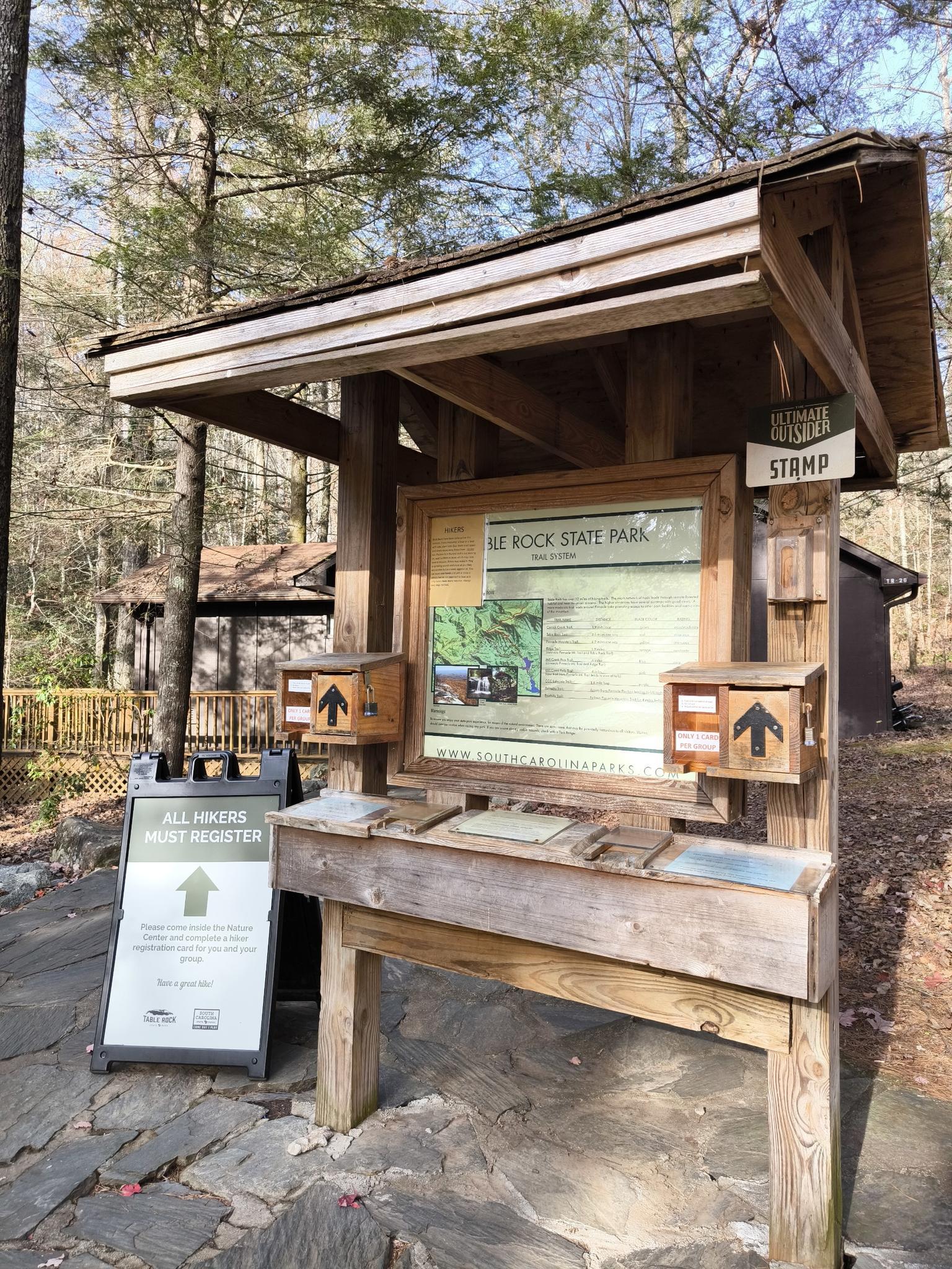 Wooden information kiosk with trail maps and signs in a sunny forest.