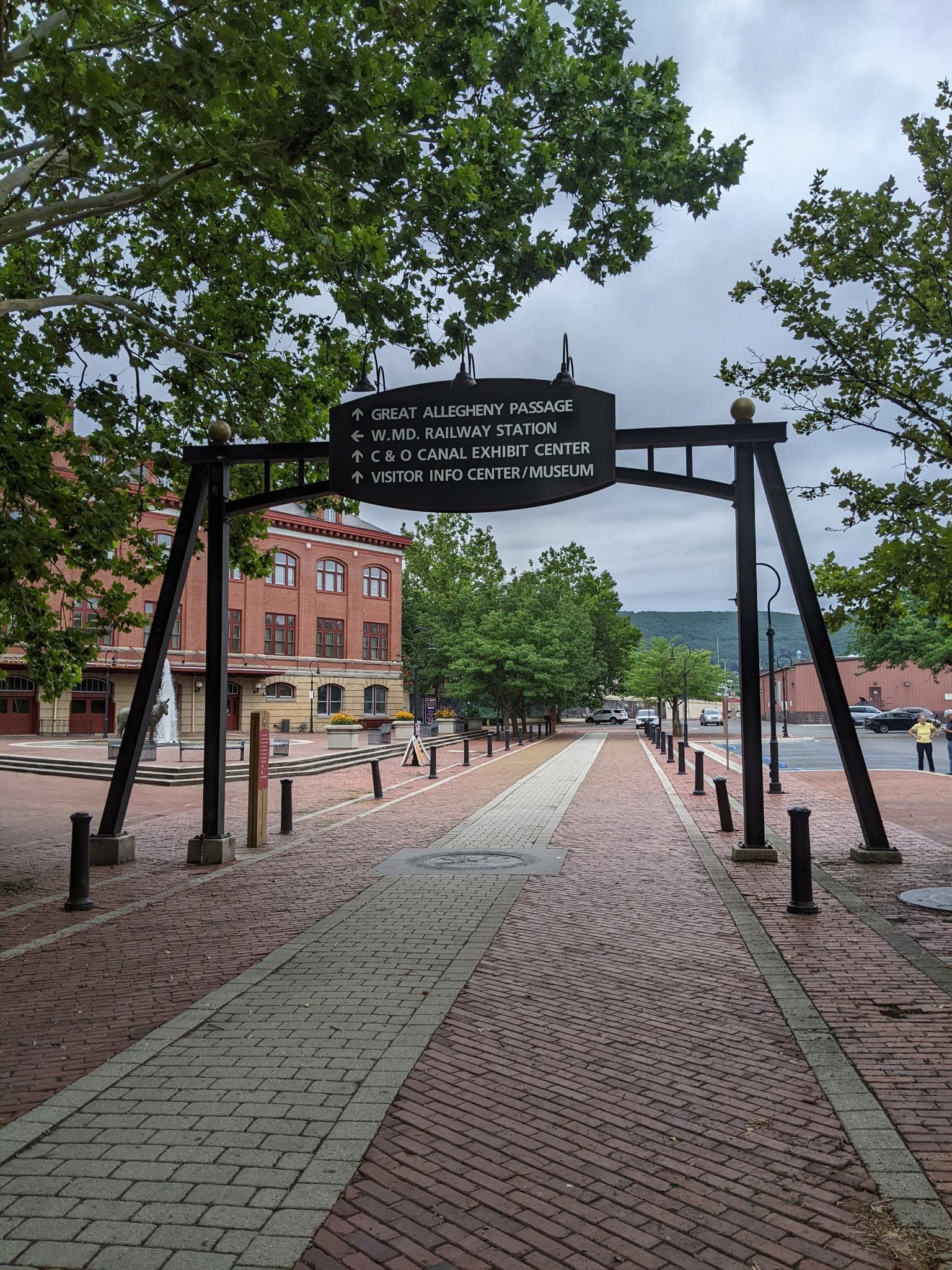 An archway sign stands over a brick path in a town square on a cloudy day.