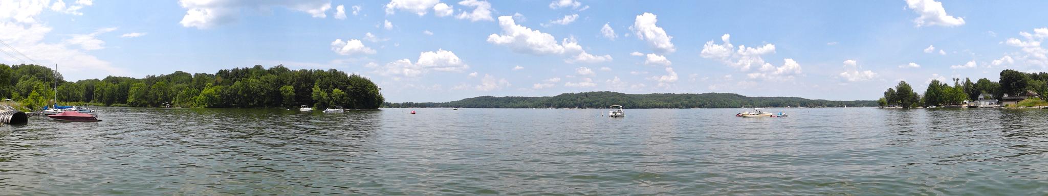 A wide panoramic view of a calm lake with distant forested shores and scattered boats under a cloudy sky.