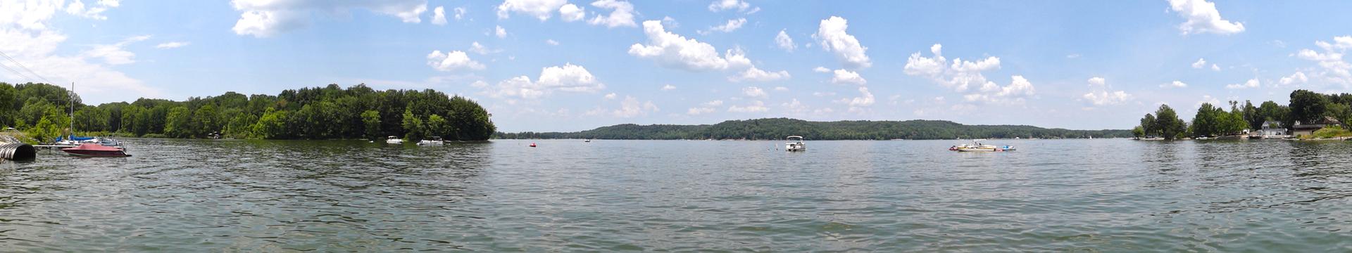 A wide panoramic view of a calm lake with distant forested shores and scattered boats under a cloudy sky.