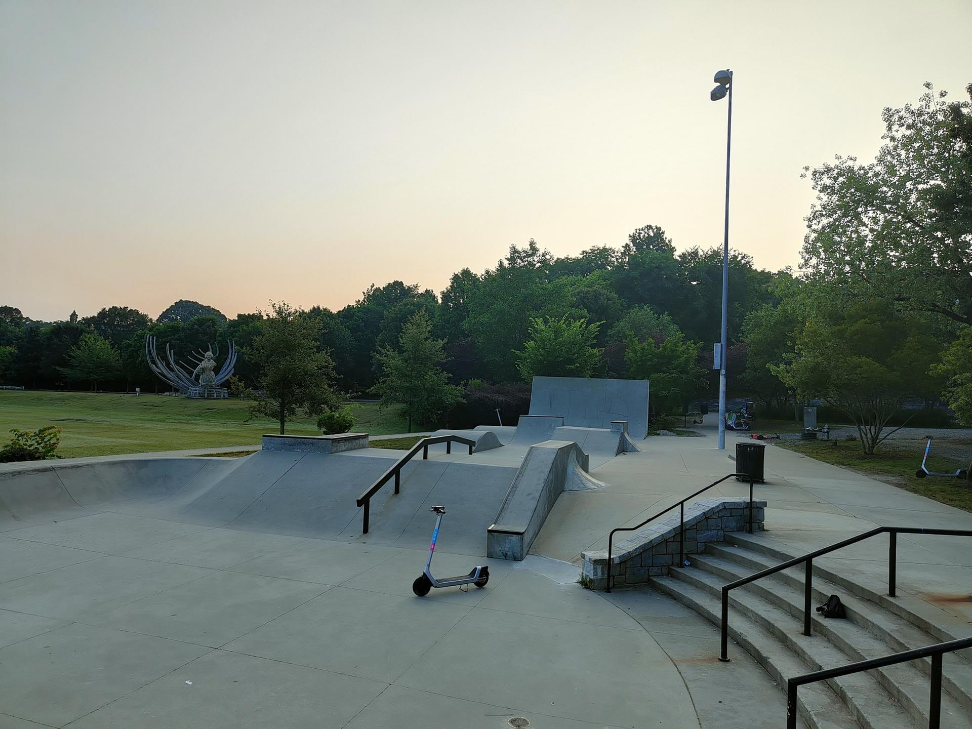 Empty skate park with ramps and a scooter at dusk, surrounded by trees.