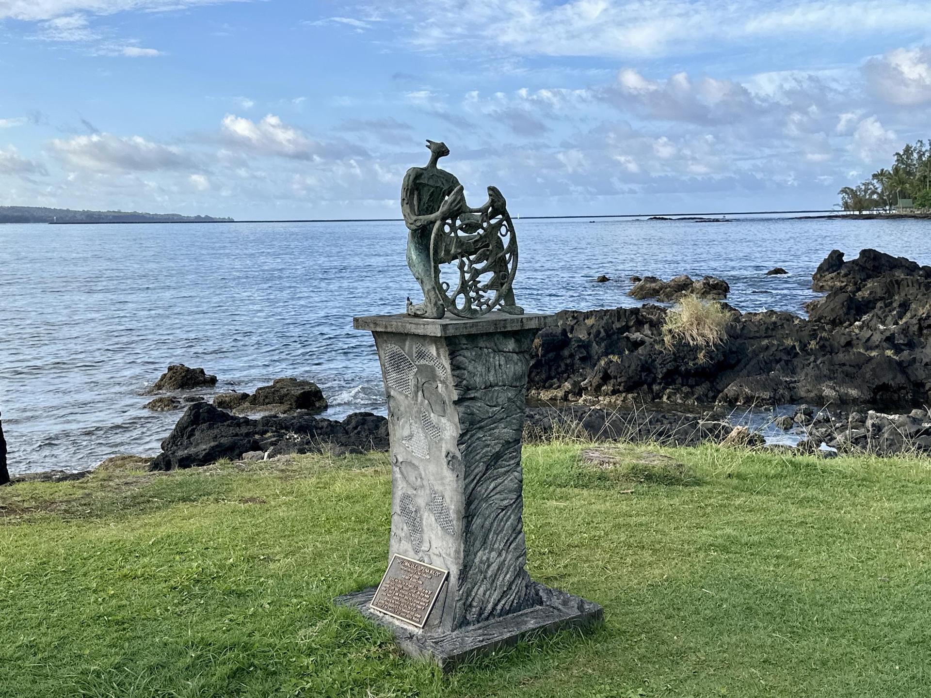Bronze statue on a pedestal by a rocky coast, calm ocean under a cloudy sky.