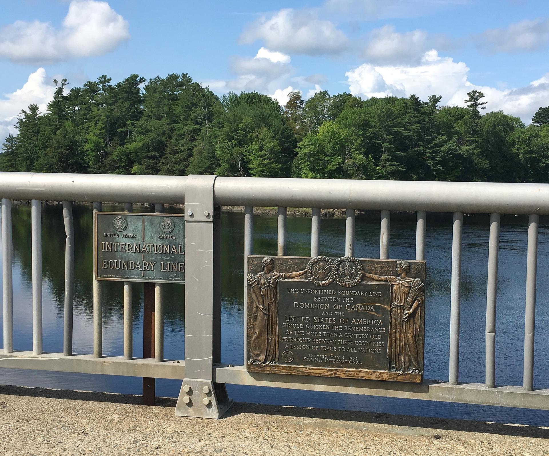 Two bronze plaques on a bridge railing overlooking water and green trees.