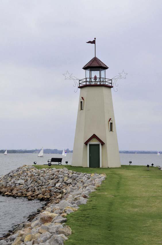 A white lighthouse stands on a rocky shore with sailboats in the distance.