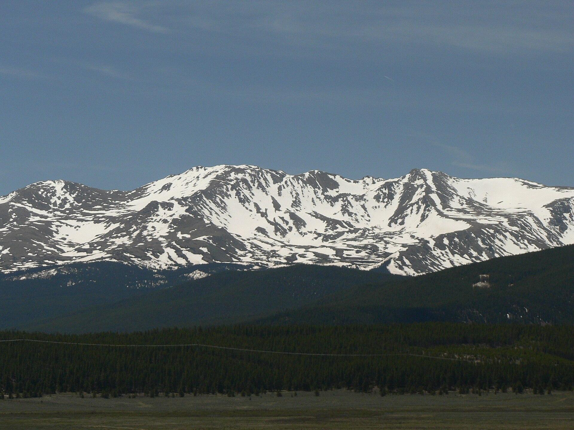 Distant snow-capped mountains rise above a dark green forest under a clear sky.