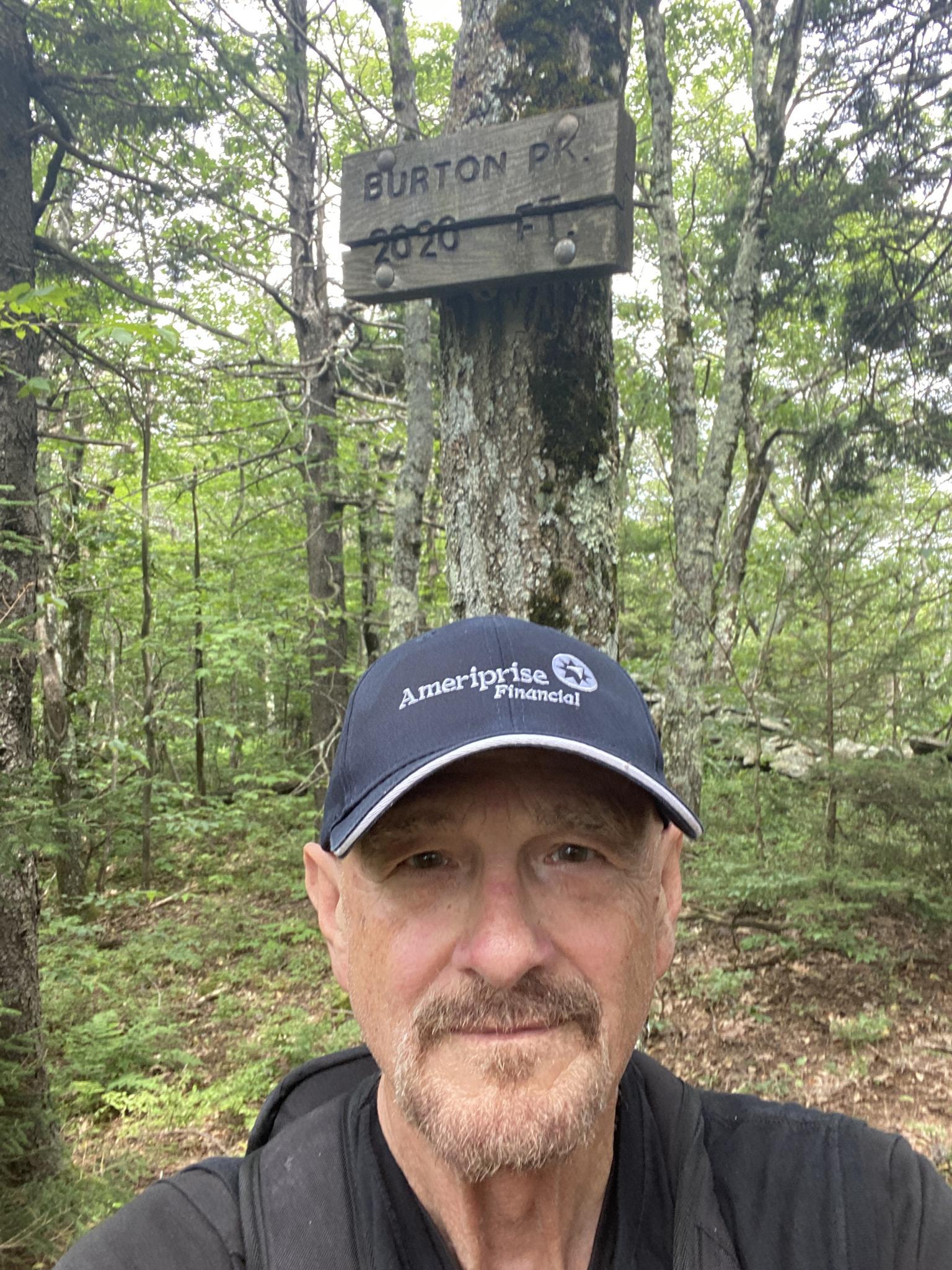 Man in a cap smiles, posing with a wooden marker in a green forest.