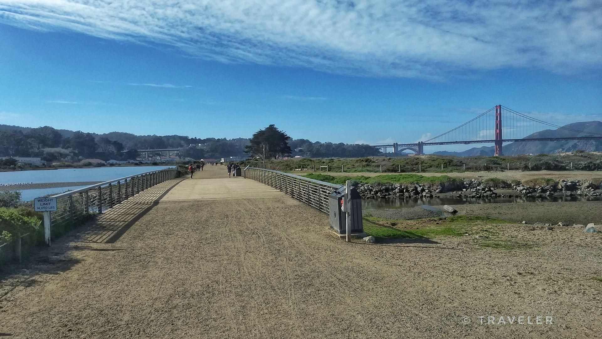 People walk on a sunny pathway leading to a large red bridge. - Golden Gate Vista Point – Marina Yacht harbor ⛵ loop from San Francisco Maritime National Historical Park