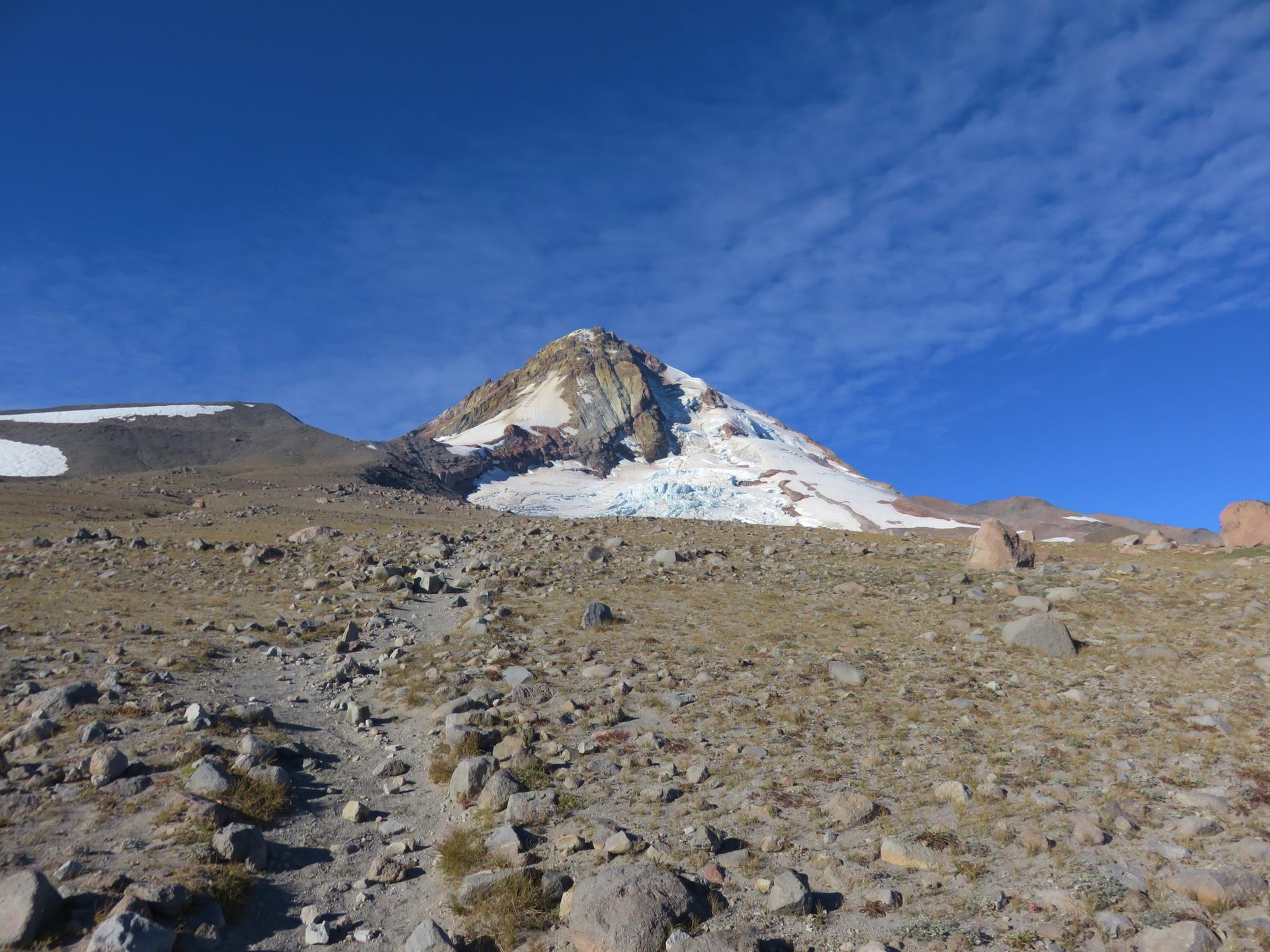 Bright daylight illuminates a rocky trail ascending towards a glacier-covered mountain.