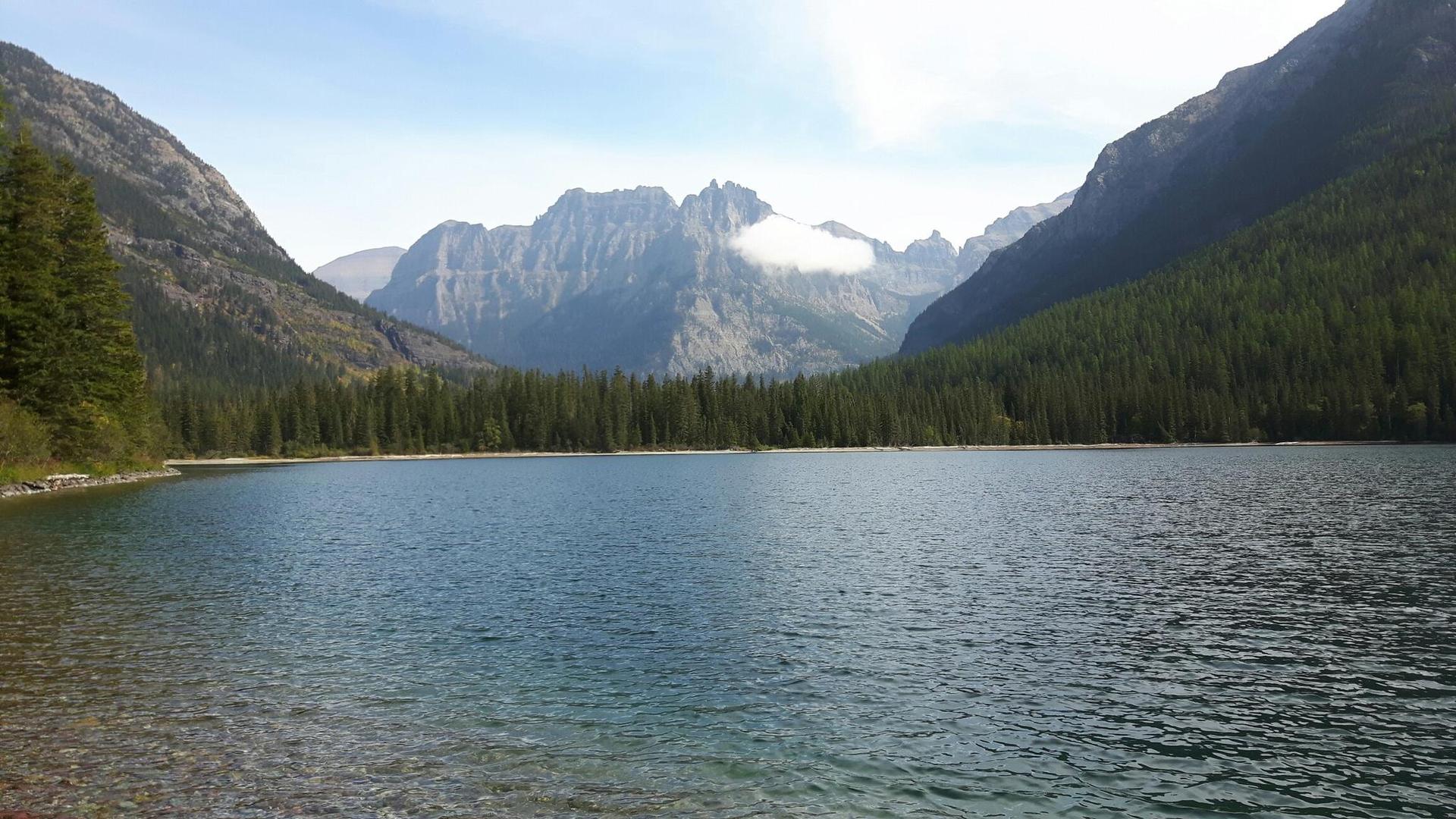 Serene lake with clear water, surrounded by forested mountains under a bright sky.