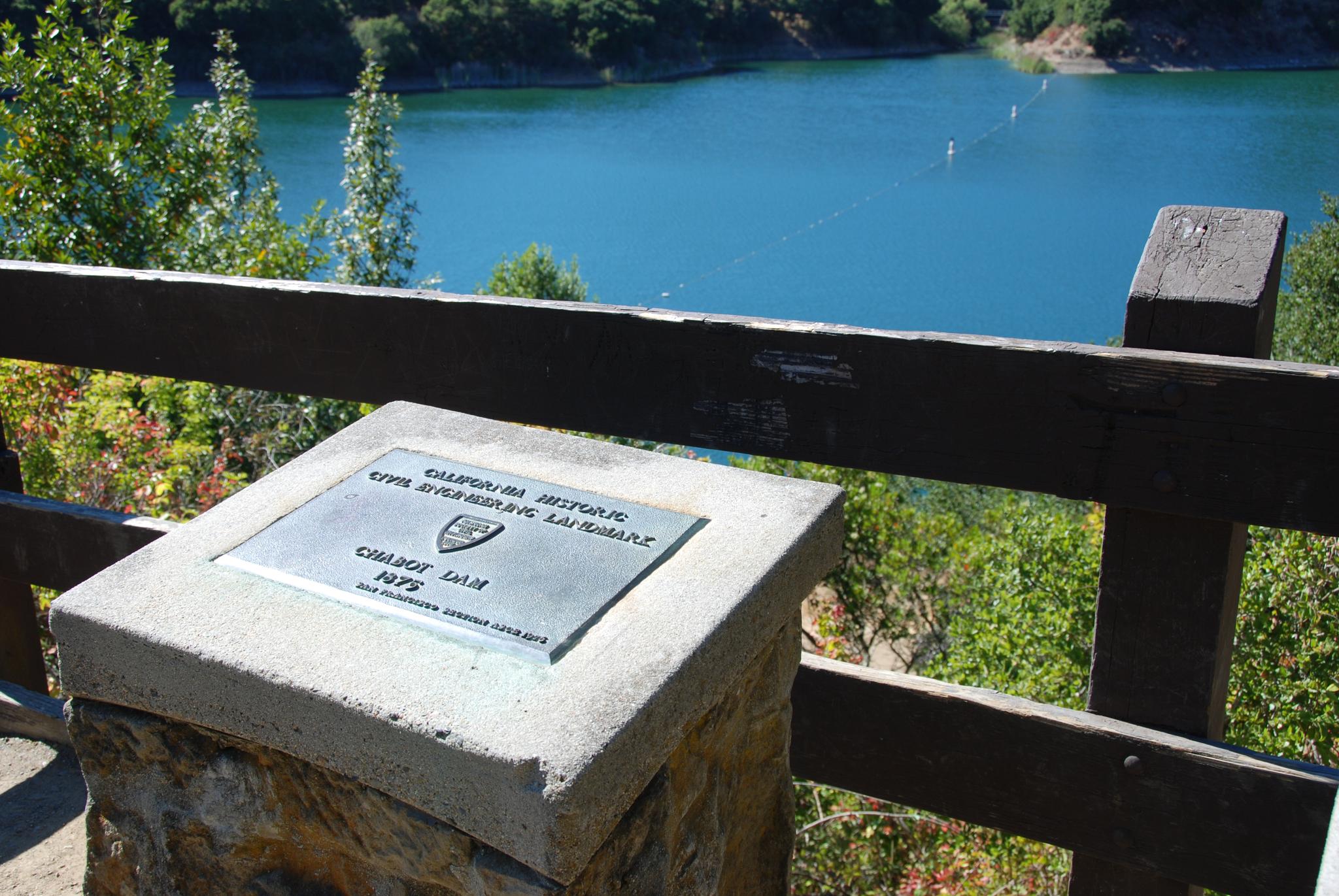 Historical marker on a stone pedestal overlooking a vibrant blue lake.