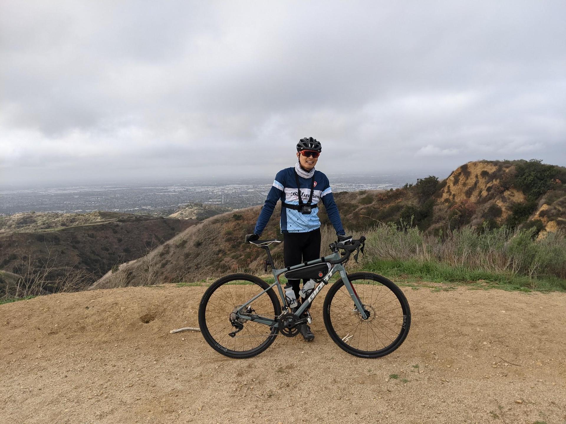 A smiling cyclist stands with their bike on a hilltop, overcast sky.