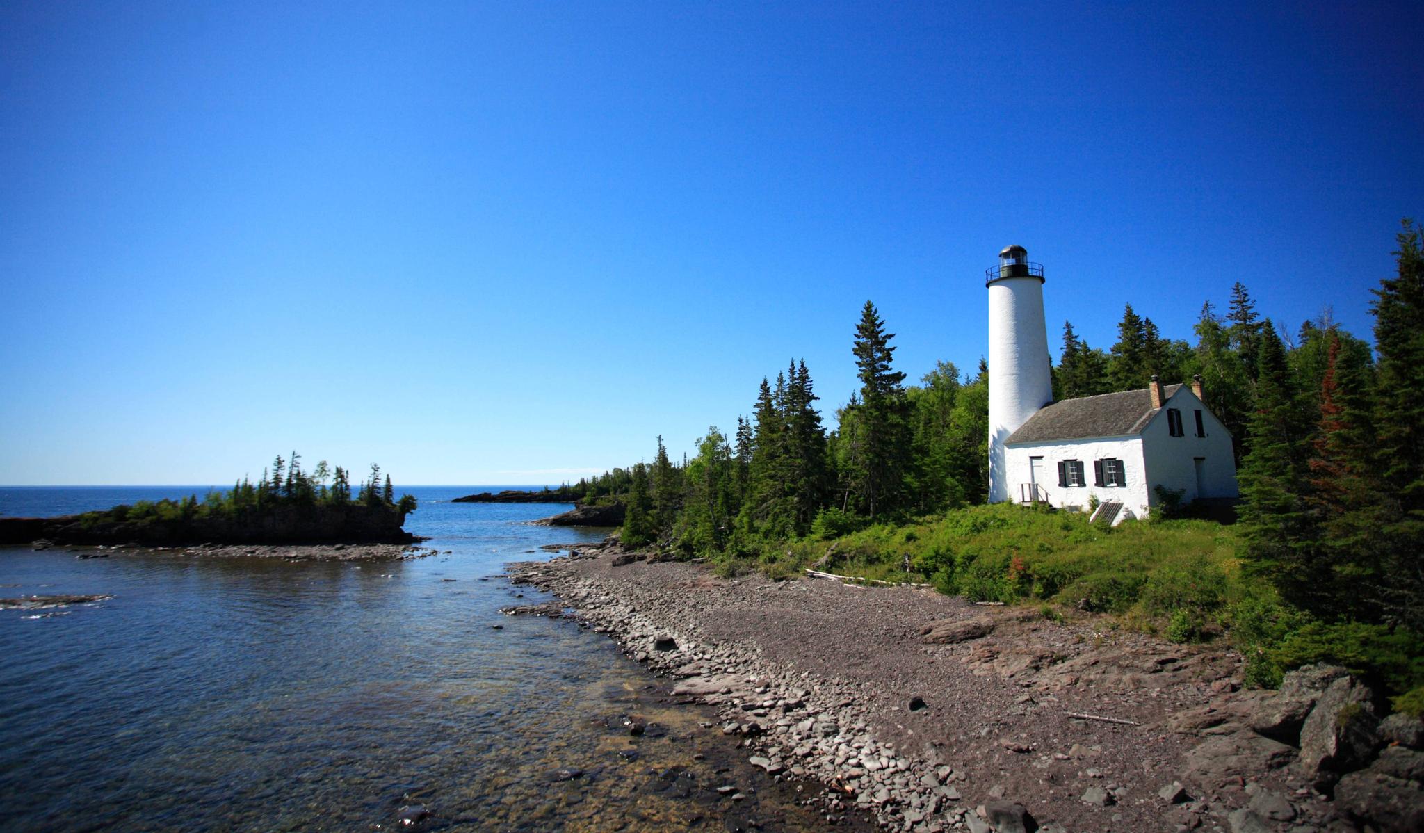 White lighthouse and house on a rocky, tree-lined coast under a clear blue sky.