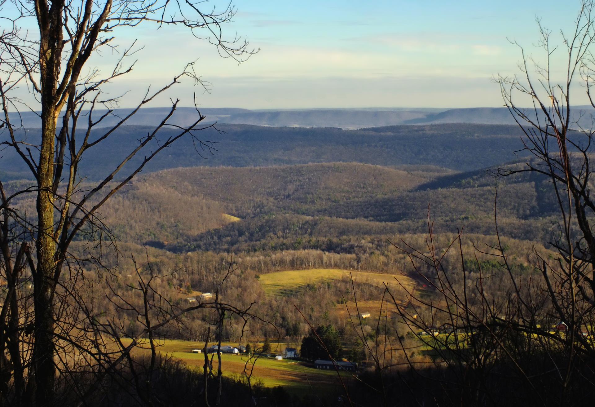 Vast mountainous landscape with valleys, distant fields, and bare trees in foreground.