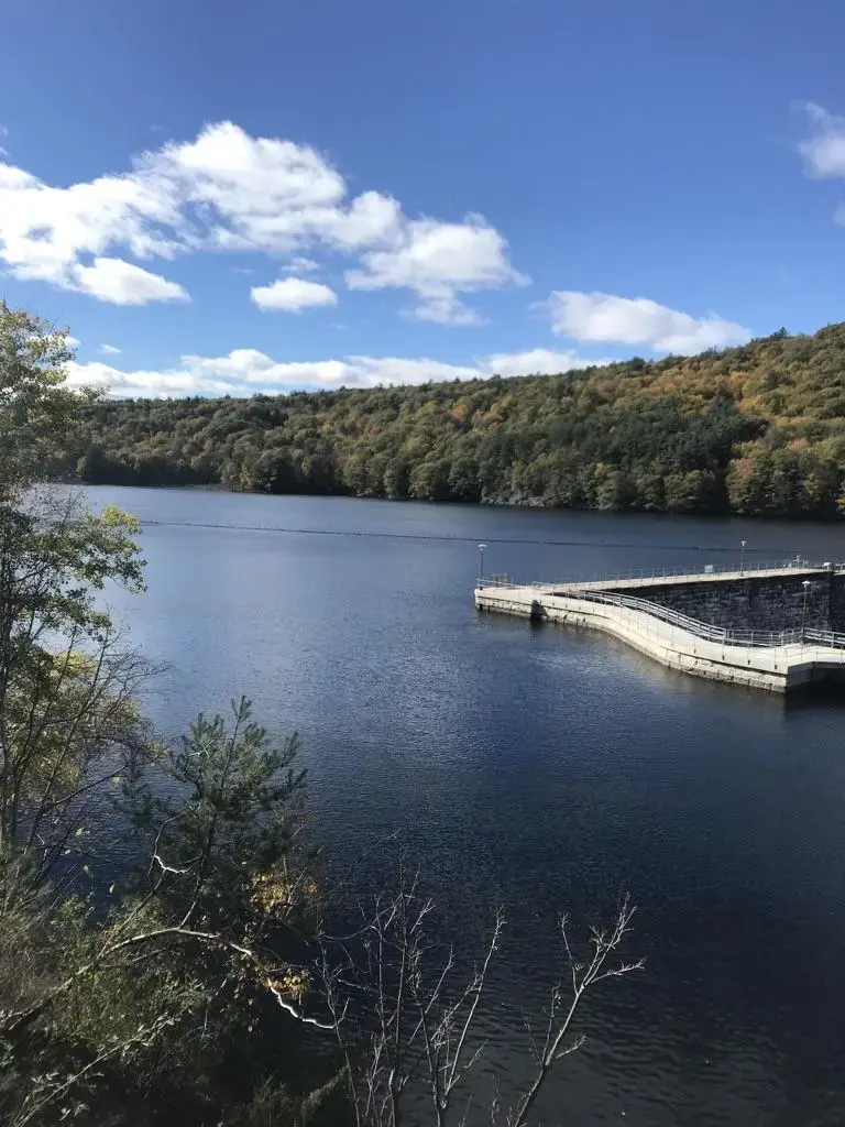 A vast lake with a concrete dam structure and forested hills under a partly cloudy sky.