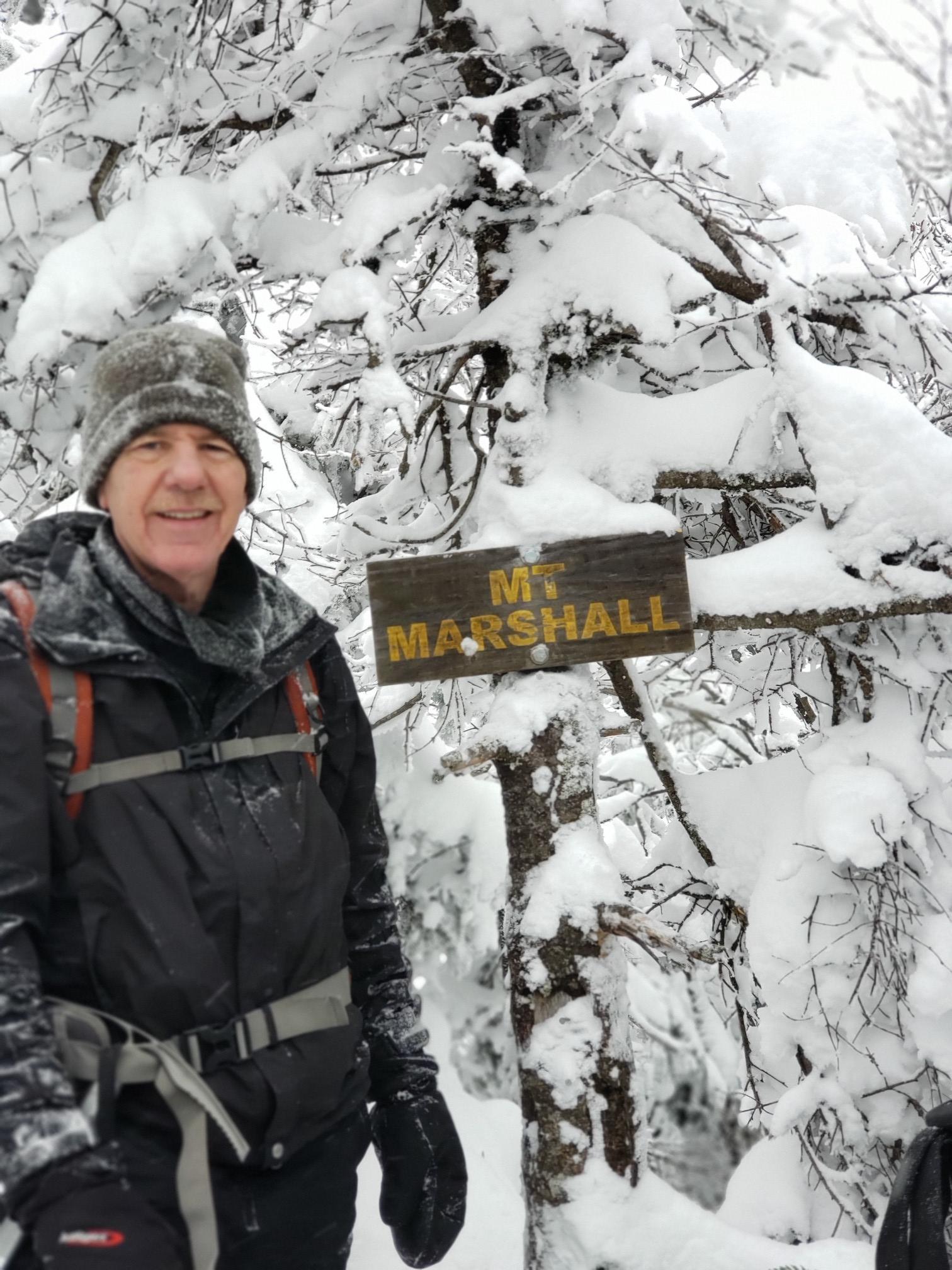 Smiling man in winter gear stands next to a snowy sign in a forest.