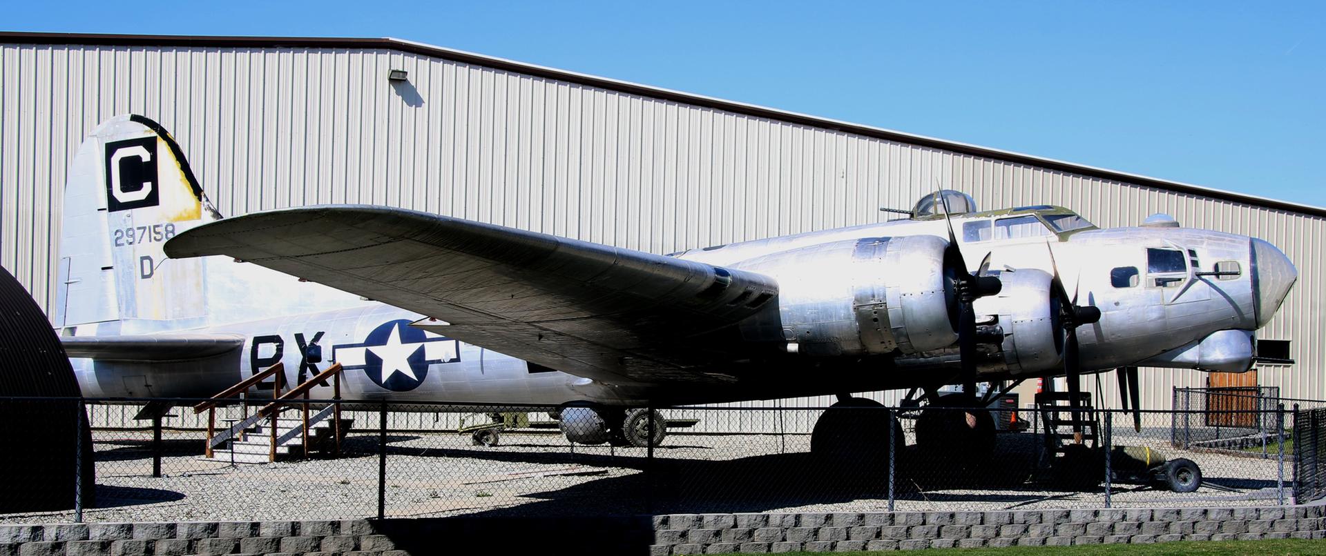 A large, silver vintage airplane parked outdoors next to a building on a sunny day.