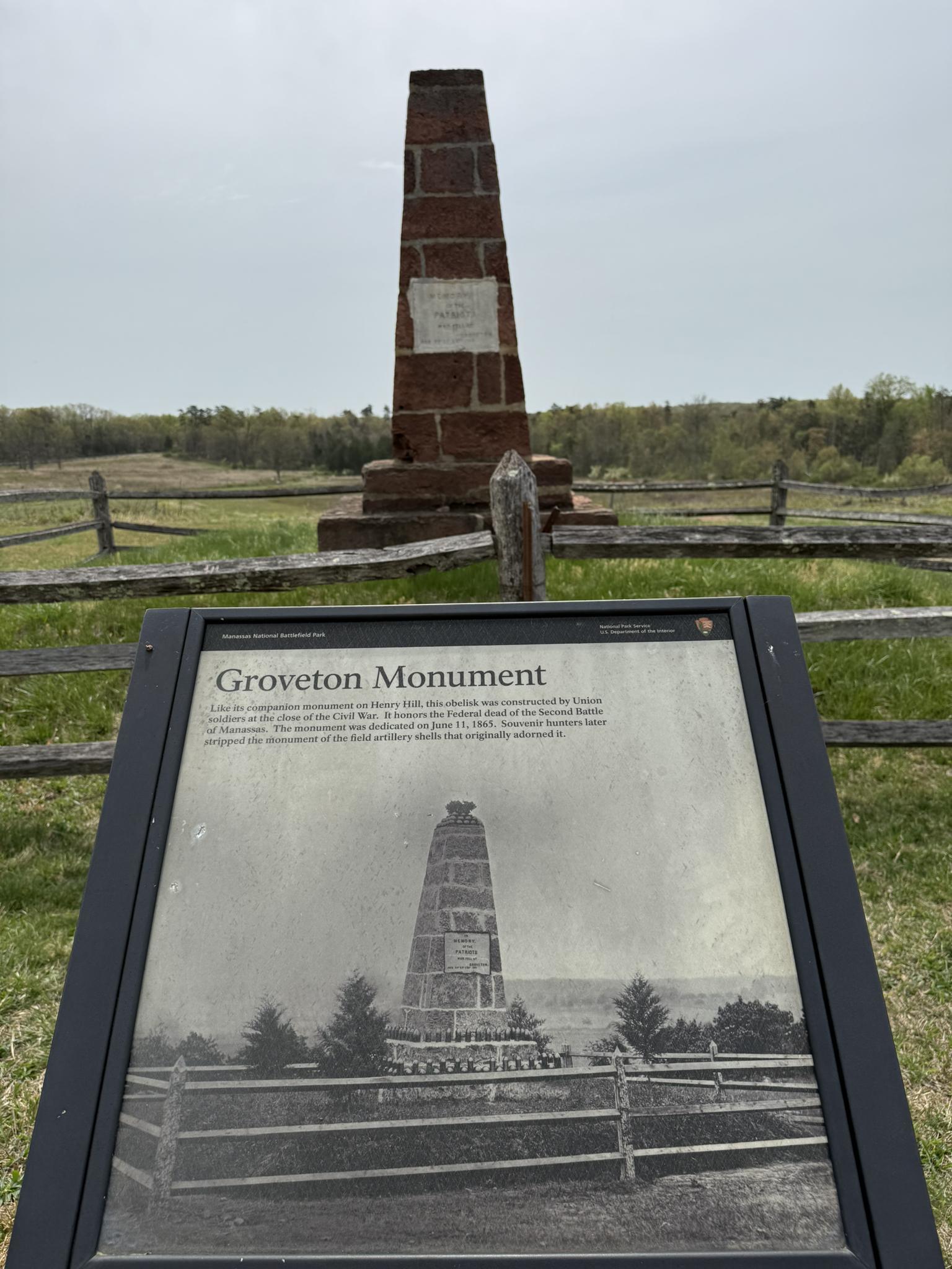 An information sign in front of a brick monument in a fenced, grassy field.