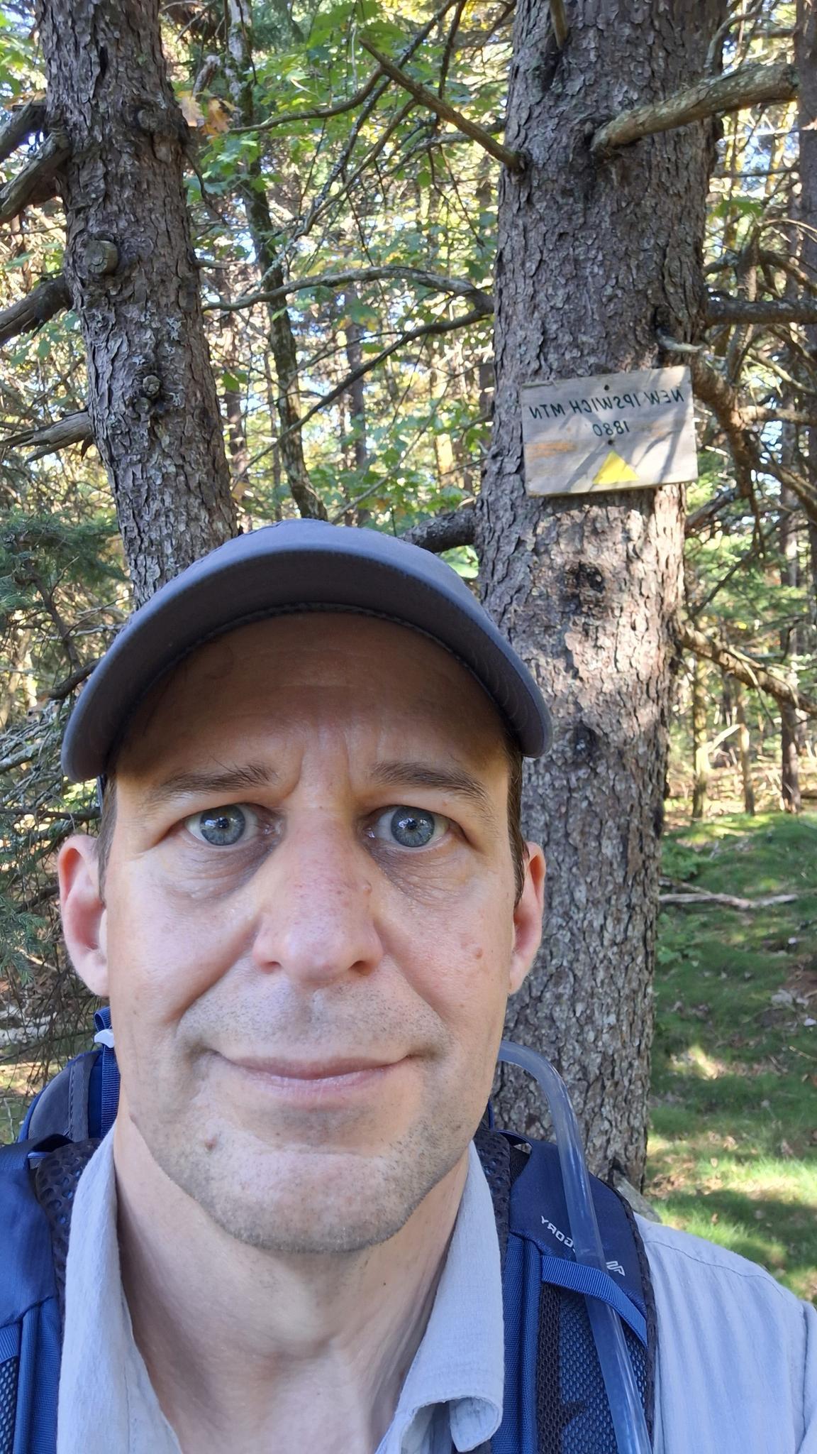 A man in a cap smiles, standing next to a tree with a sign.