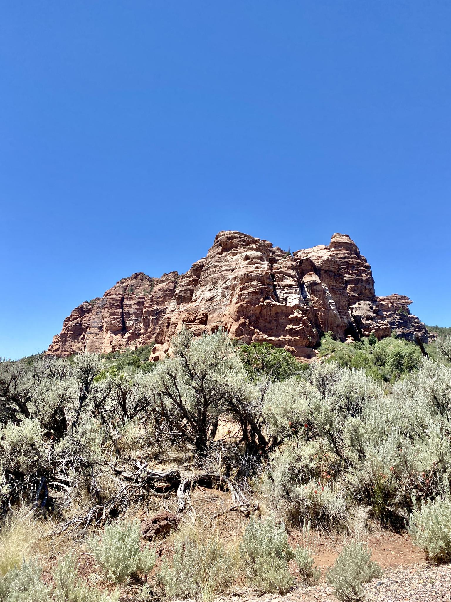 Rugged red rock formation rises above desert scrubland under a bright blue sky.