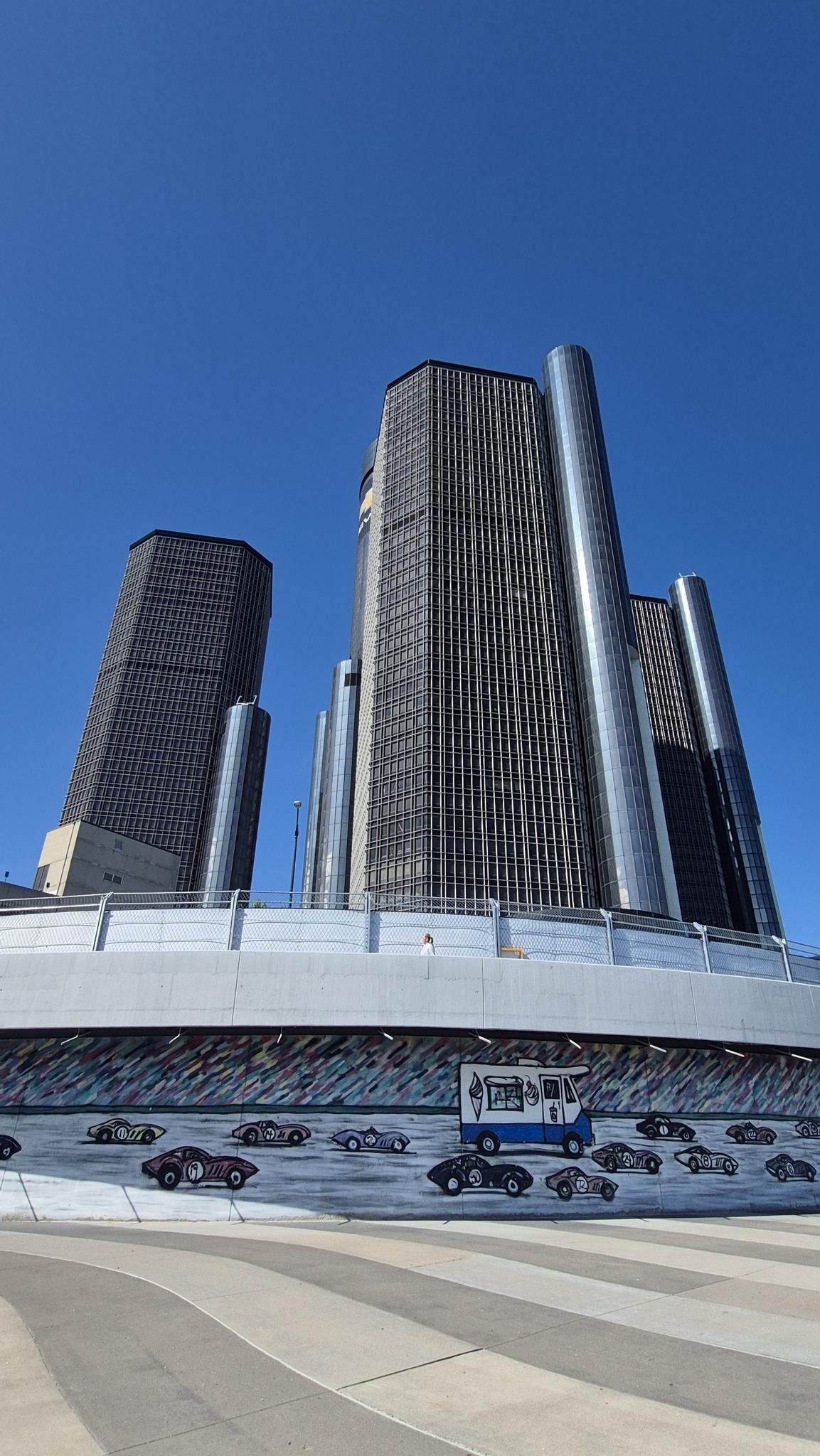 Modern skyscrapers against a blue sky, with a car mural below. - Renaissance Center – Gateway to Freedom Monument loop from Detroit