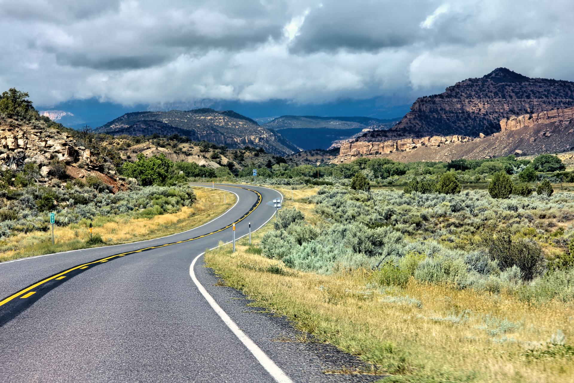 Winding road through a desert landscape under a cloudy sky, with distant mountains.