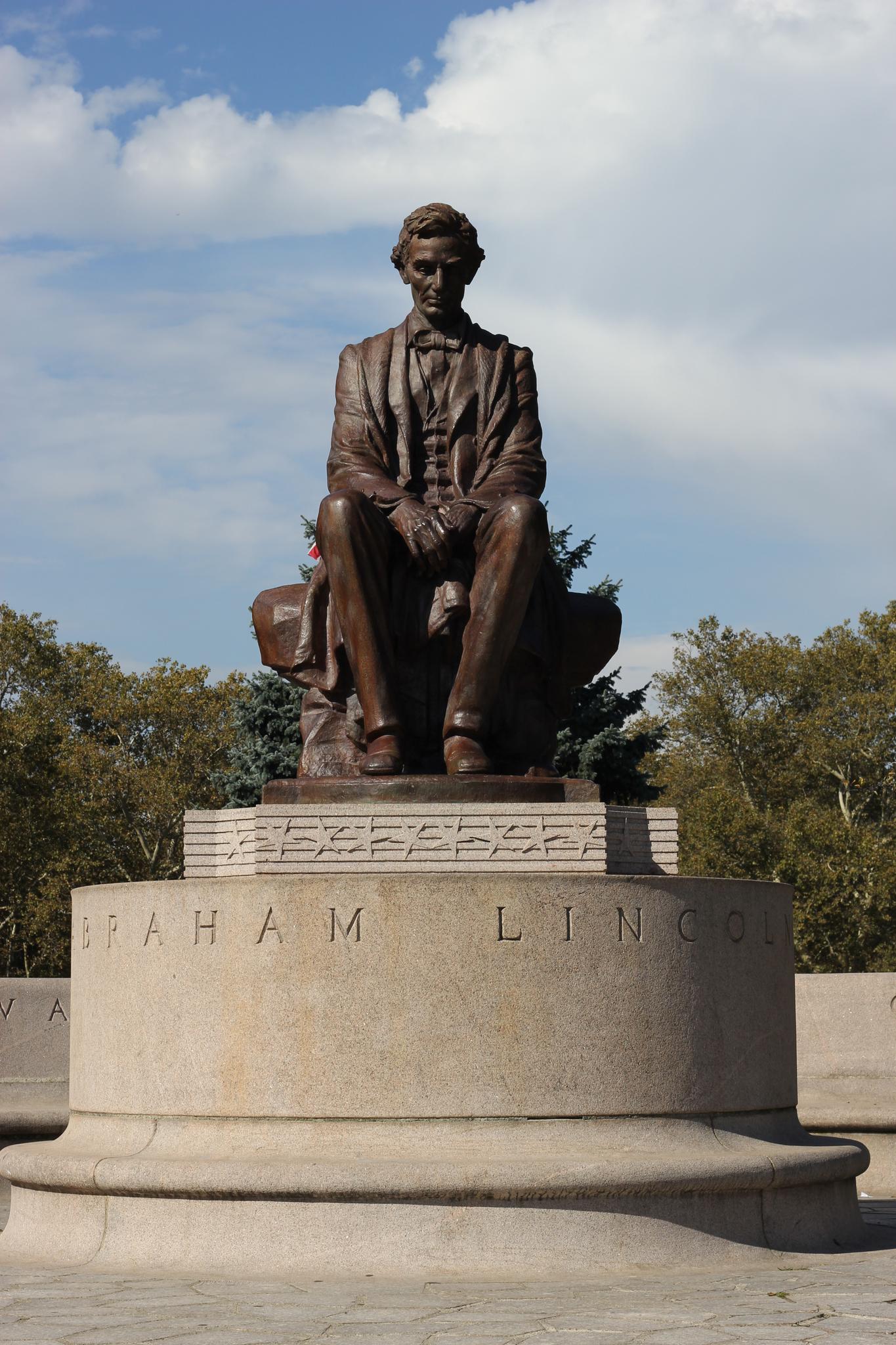 Bronze statue of a seated man on a stone pedestal, sunny day.