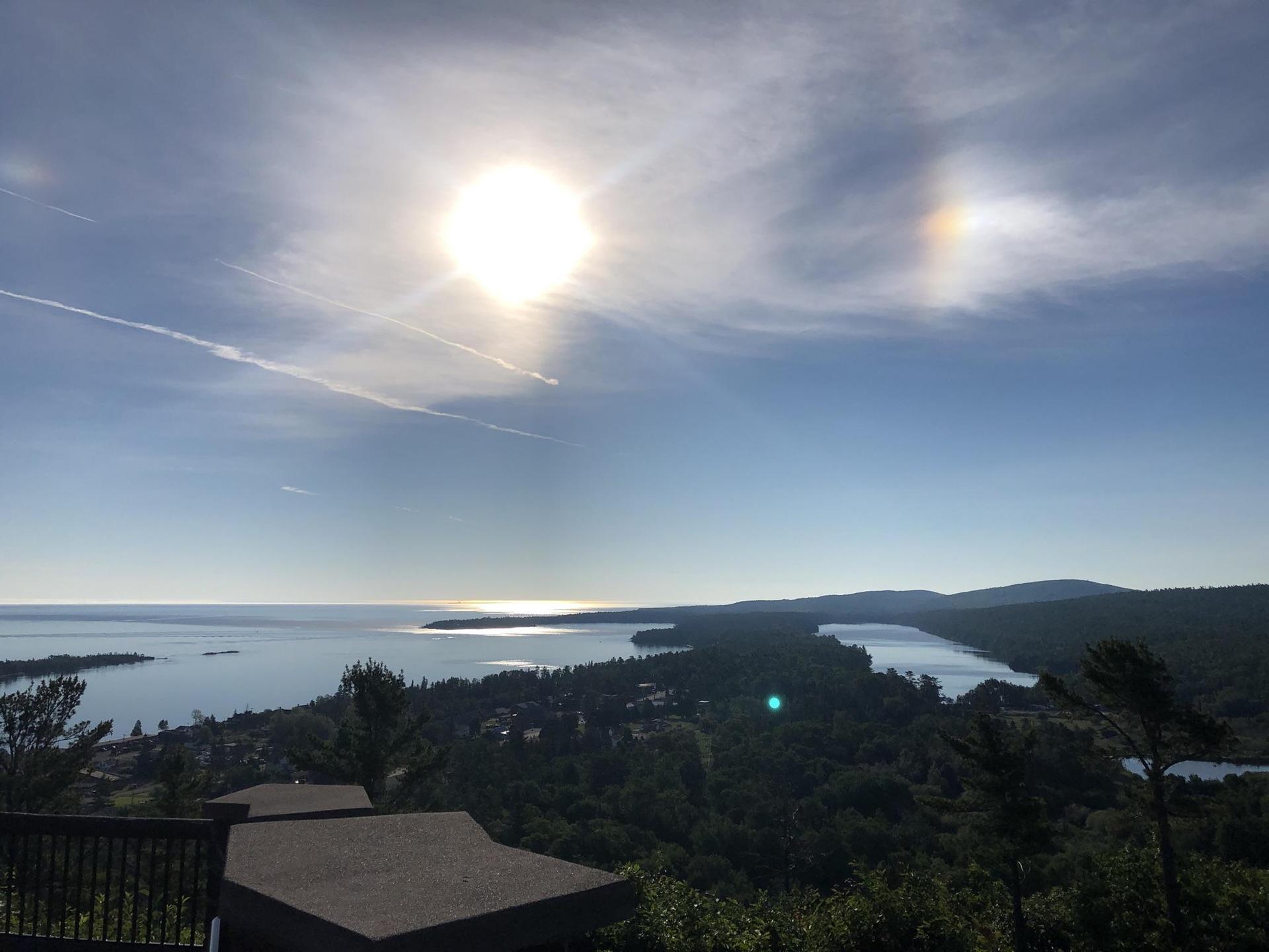 Bright sun over a large body of water, green forests, and distant hills.
