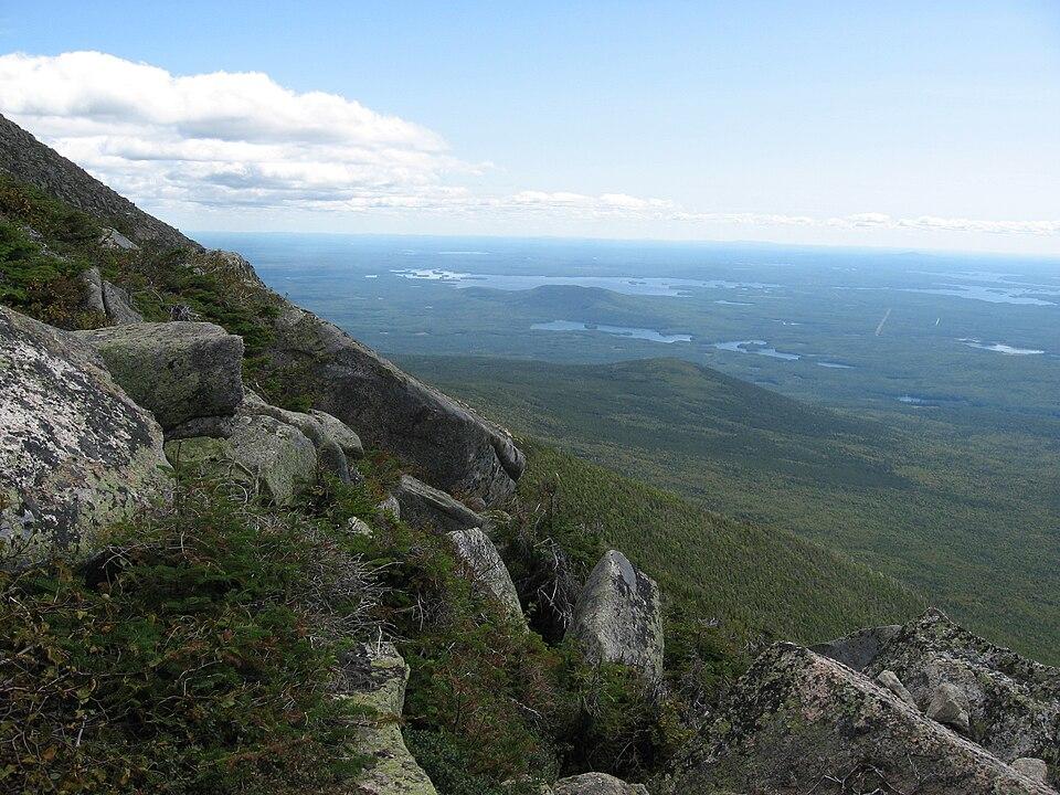 Steep rocky mountain slope overlooking a vast forested landscape with distant lakes.