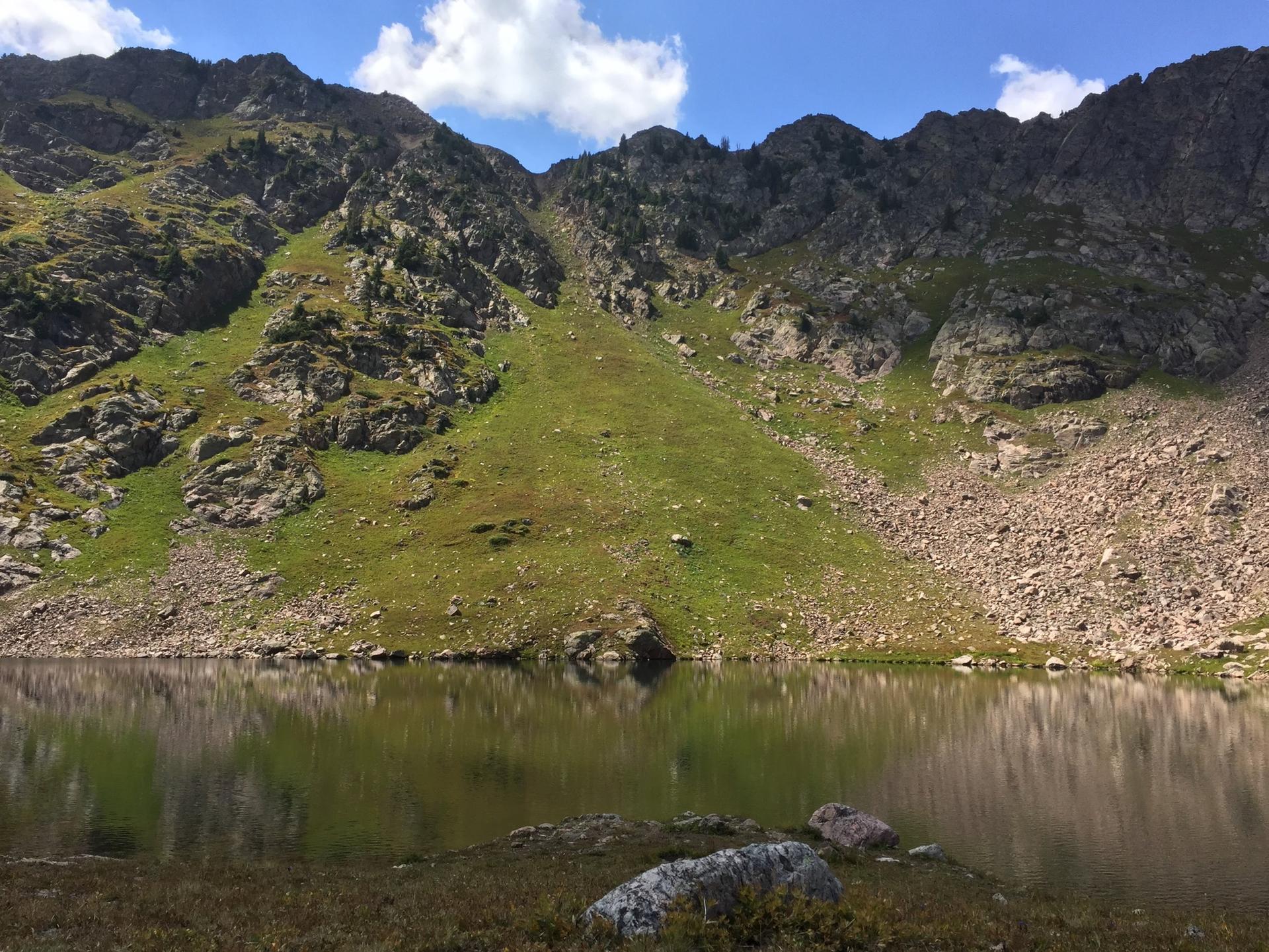 Serene mountain lake reflecting rocky, green slopes under a blue sky.