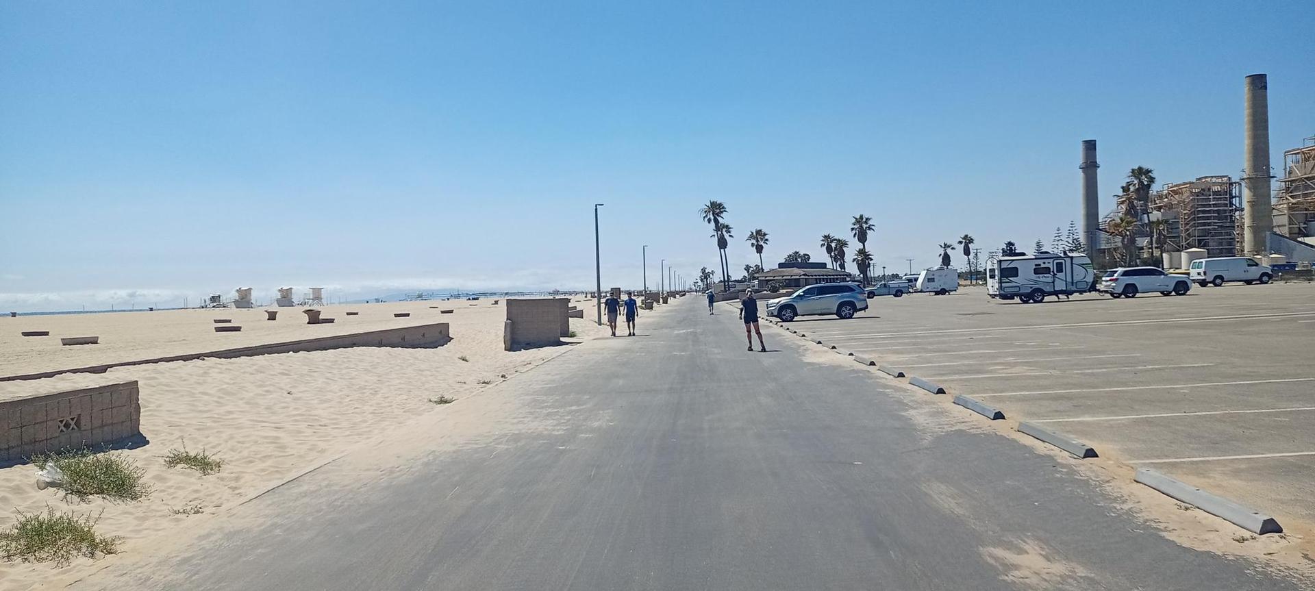 Paved path along a sunny beach with people, parked vehicles, and palm trees.