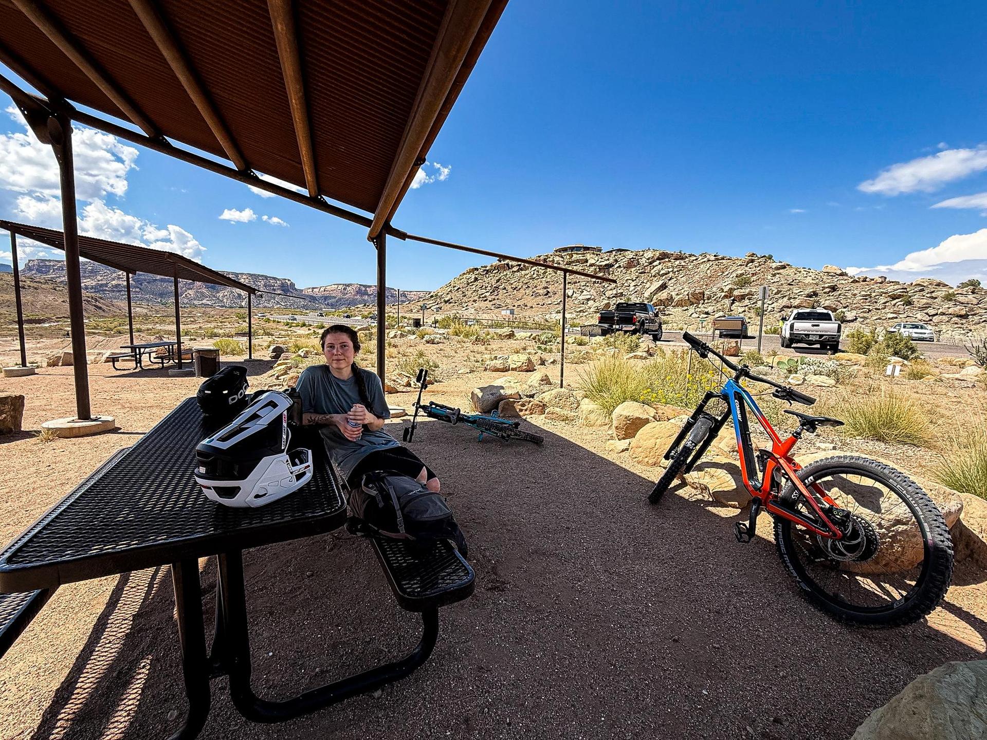 Person rests on picnic bench under shelter, mountain bike nearby.