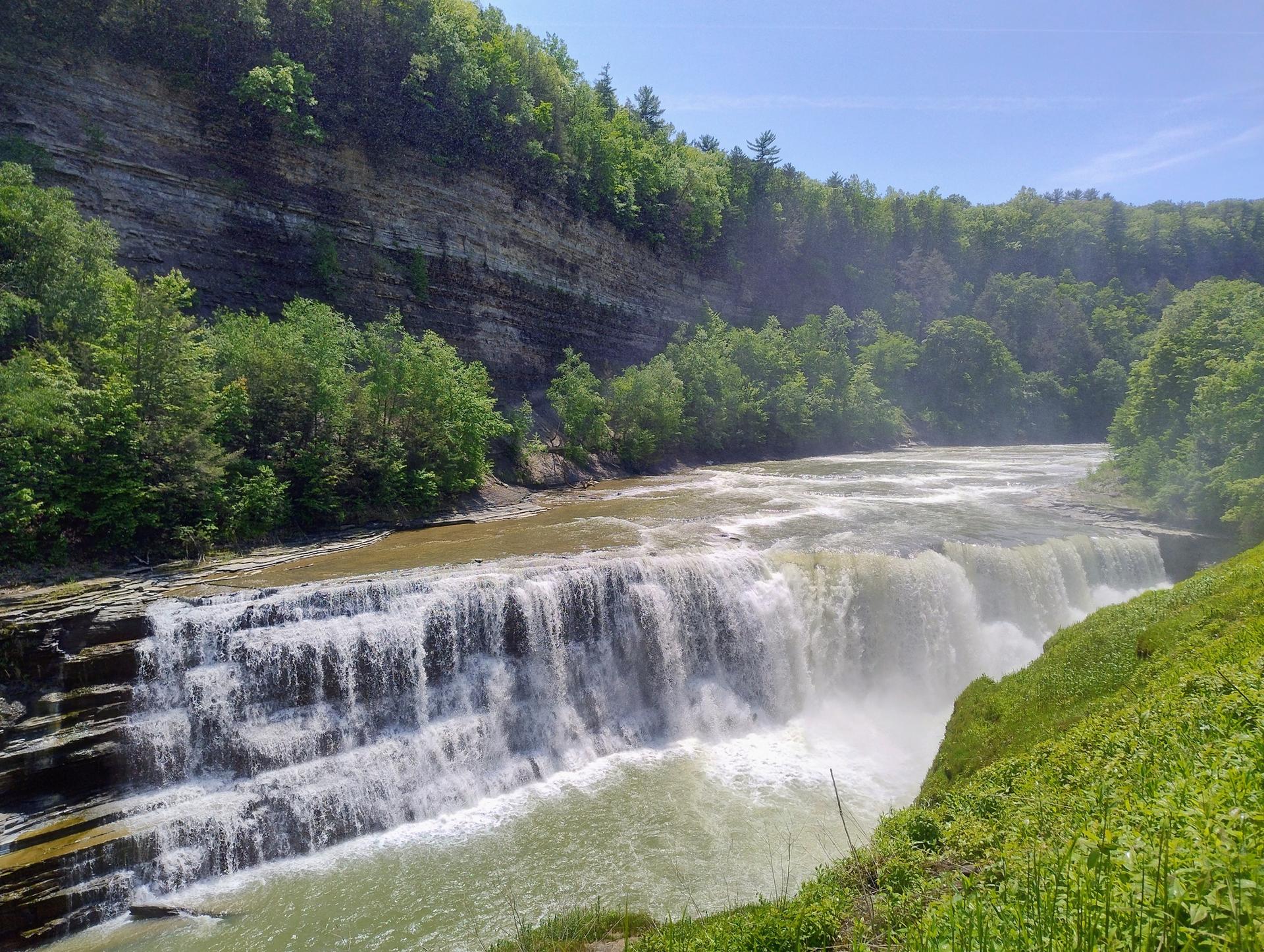 A wide, multi-tiered waterfall cascades into a river surrounded by green forests.
