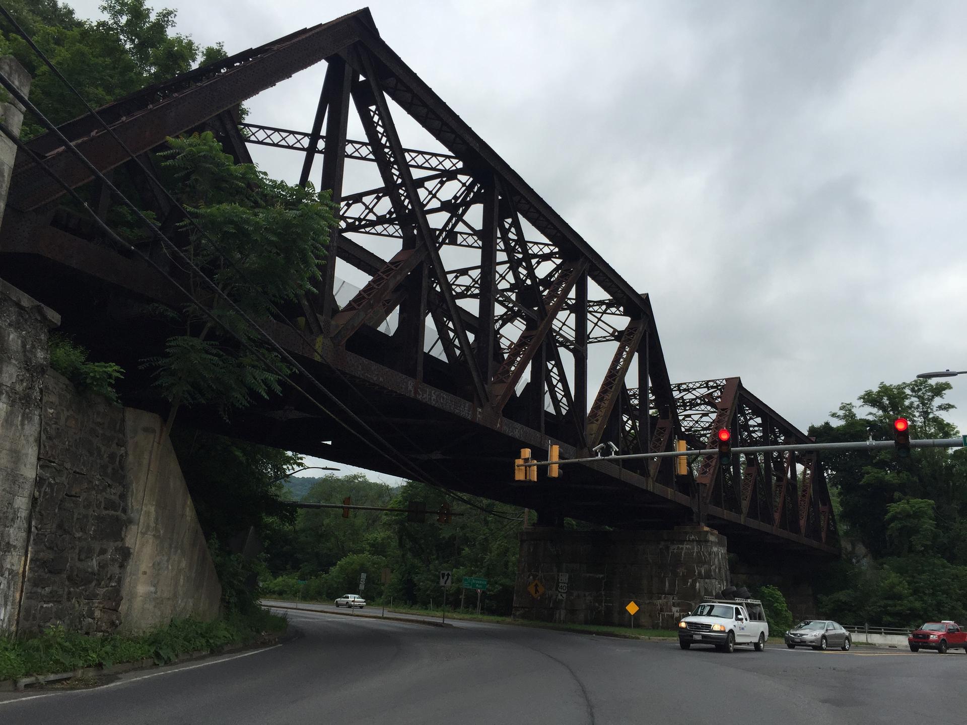 Large rusty truss bridge spans over a road with cars on an overcast day.