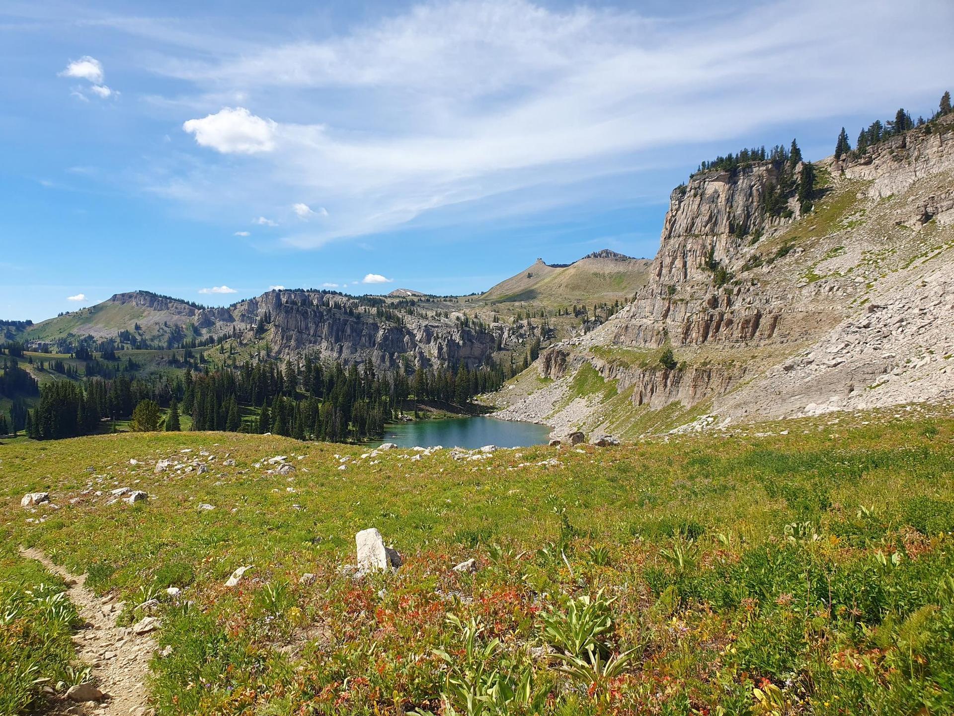 A trail overlooks a serene blue lake nestled among rocky cliffs and green hills.