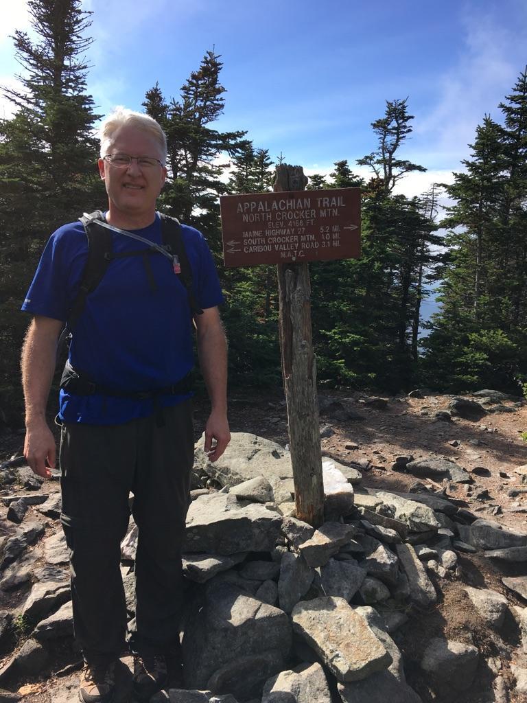 Man in blue shirt standing next to a wooden trail sign.