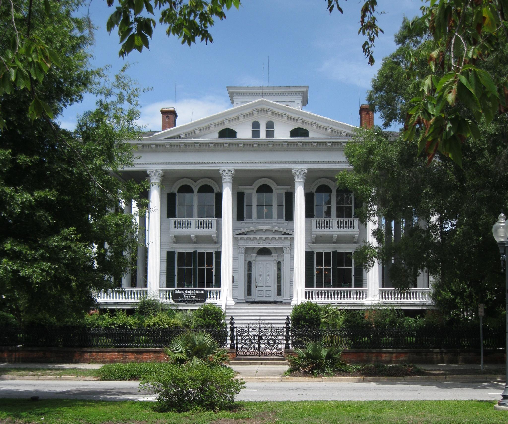 Grand white mansion with columns and balconies, surrounded by green trees.