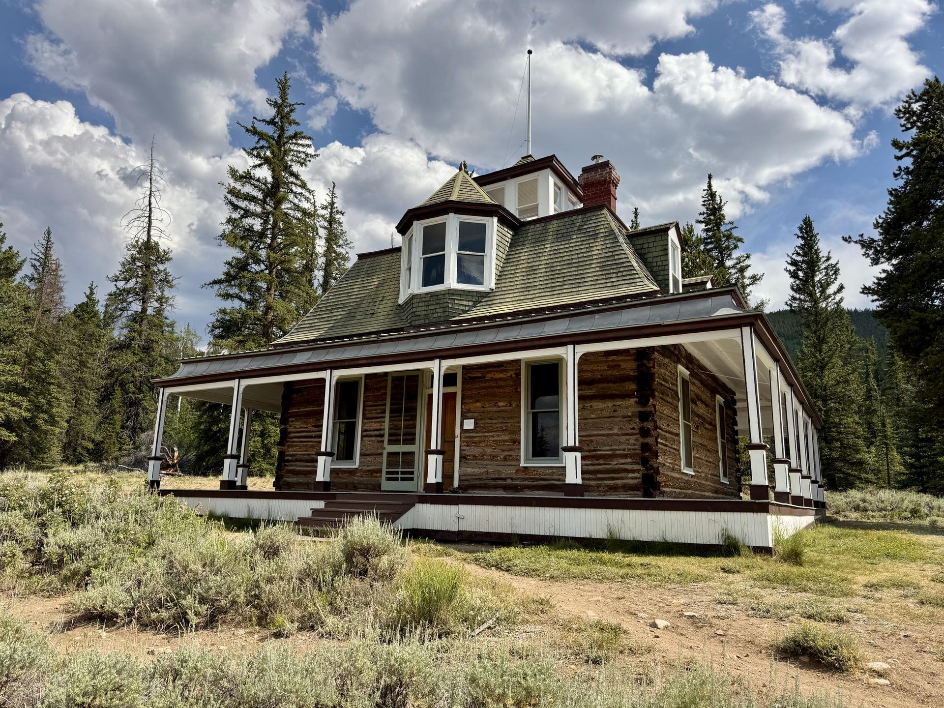 Historic wooden house with a porch, surrounded by pine trees under a cloudy sky.