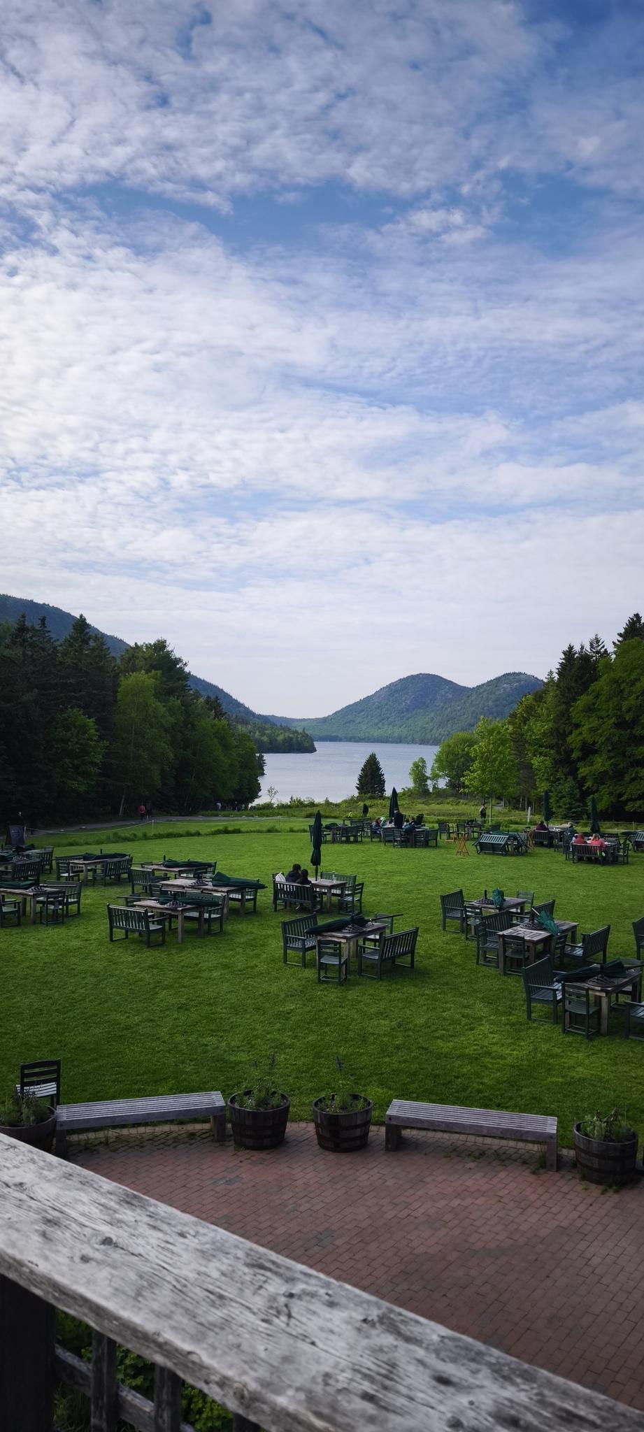 Grassy lawn with outdoor tables, lake, and mountains under cloudy sky.