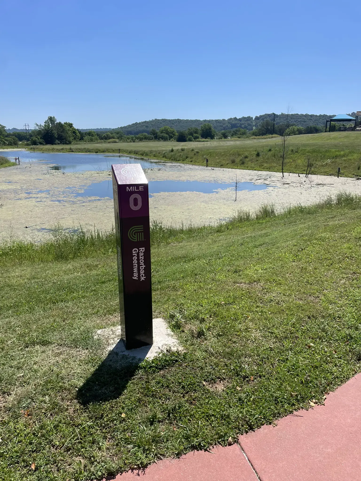 A "Mile 0" sign stands beside a green pond under a bright blue sky.