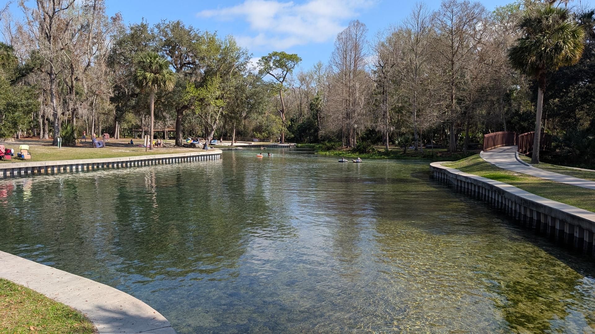 Clear river with people swimming, surrounded by trees and grassy banks.