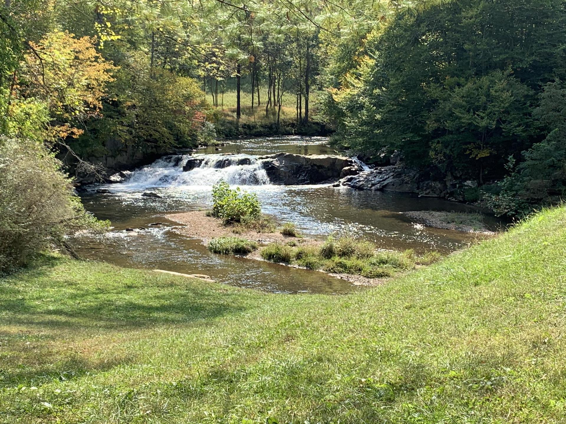 Waterfall in a sunny forest with a wide river and green banks.