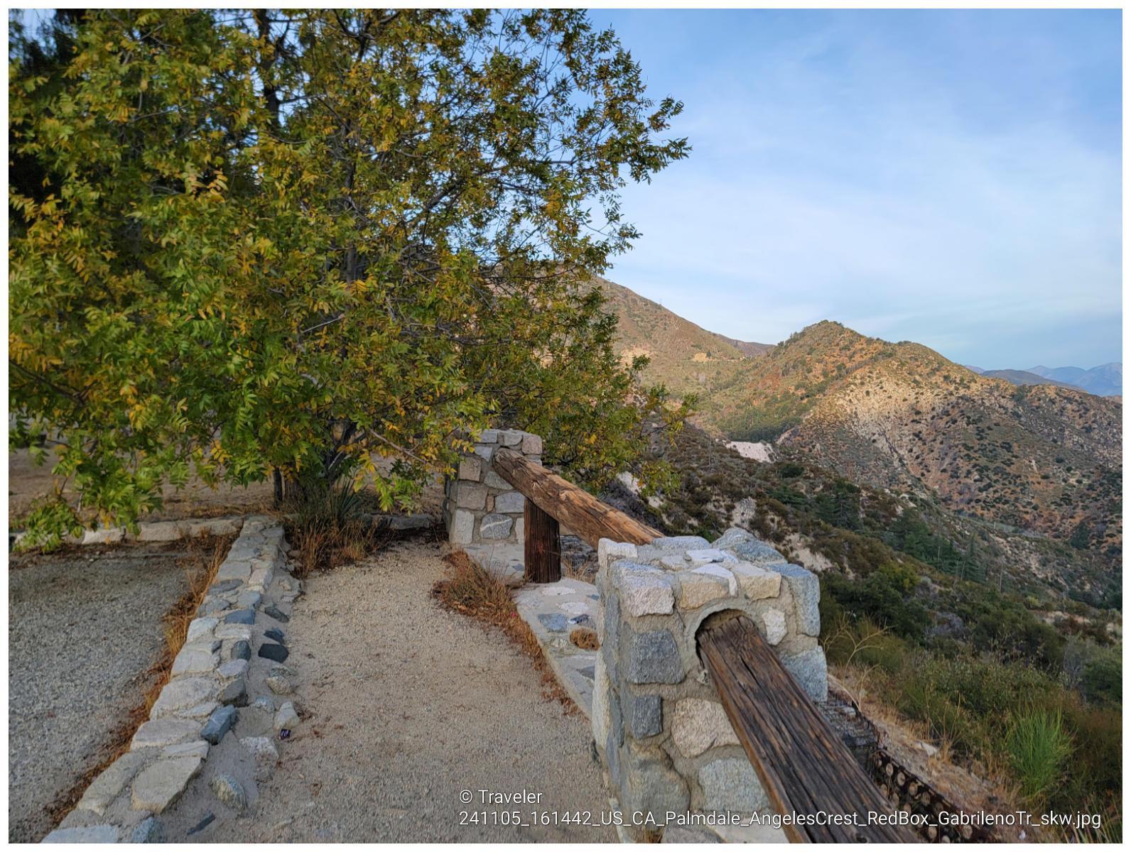 Stone and wood railing on a dirt path overlooking a vast, arid mountain landscape.