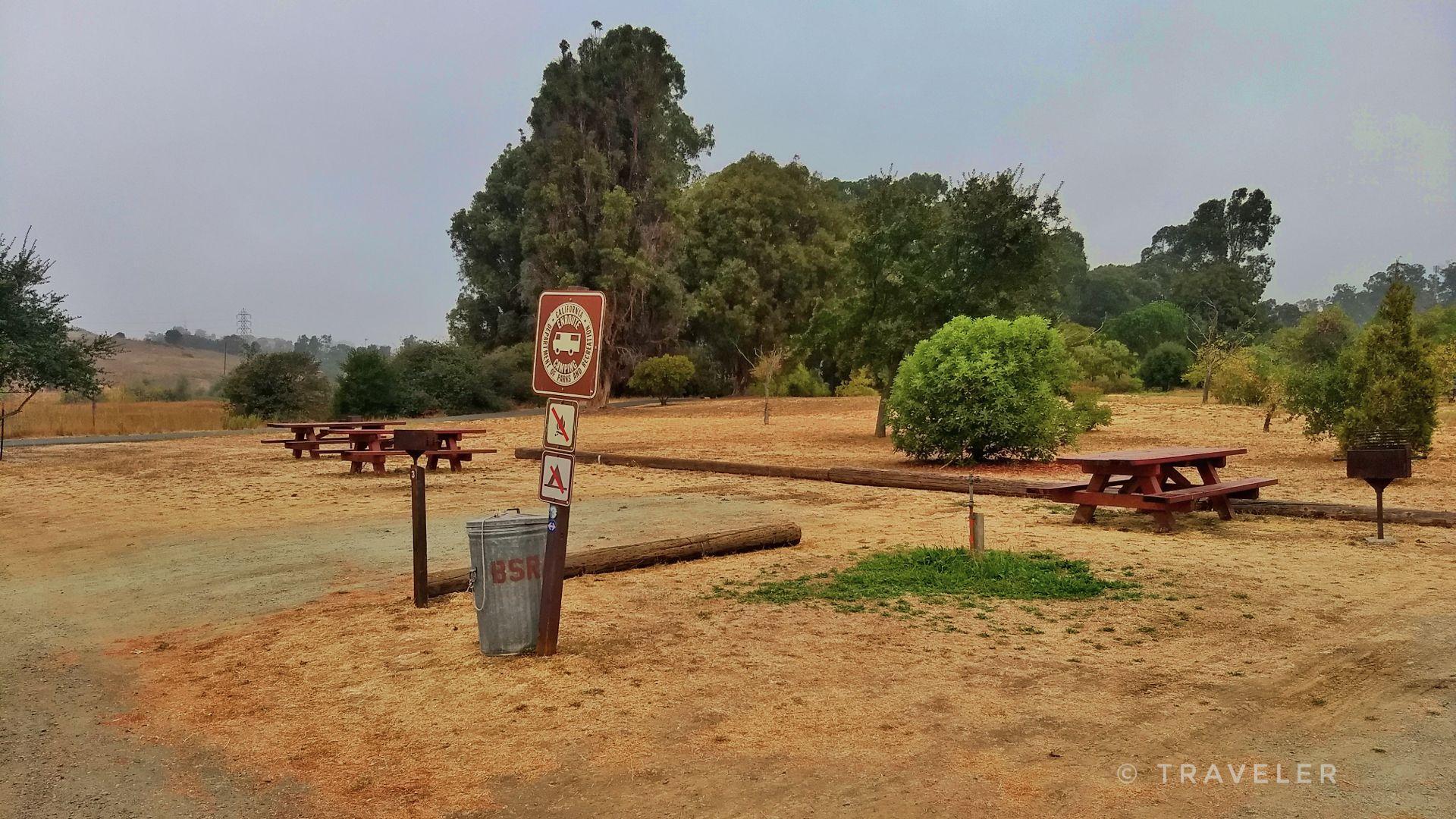 Picnic tables and signs in a dusty, wooded area under an an overcast sky.