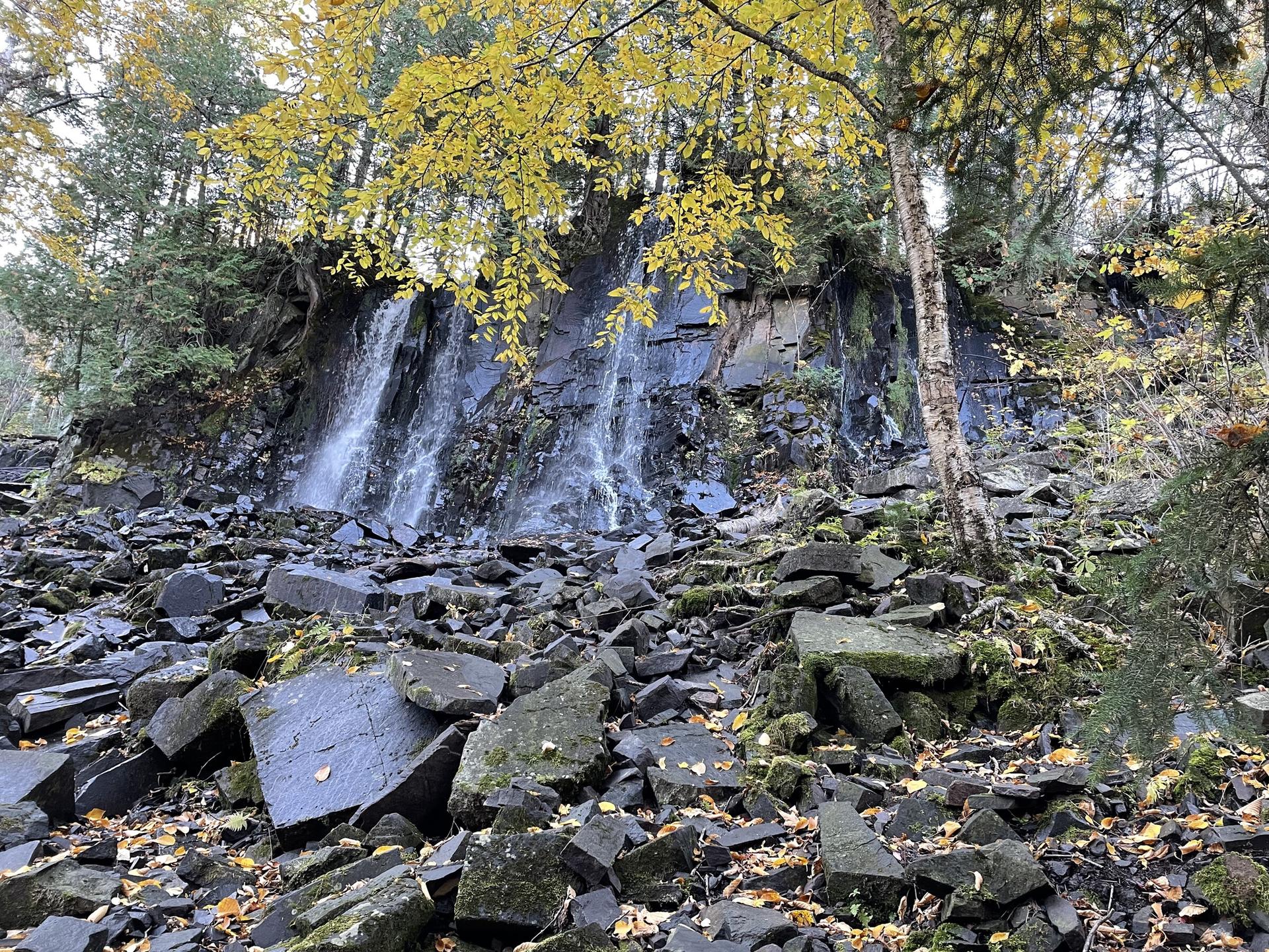 Multiple waterfalls cascade down a rocky cliff with autumn trees overhead.