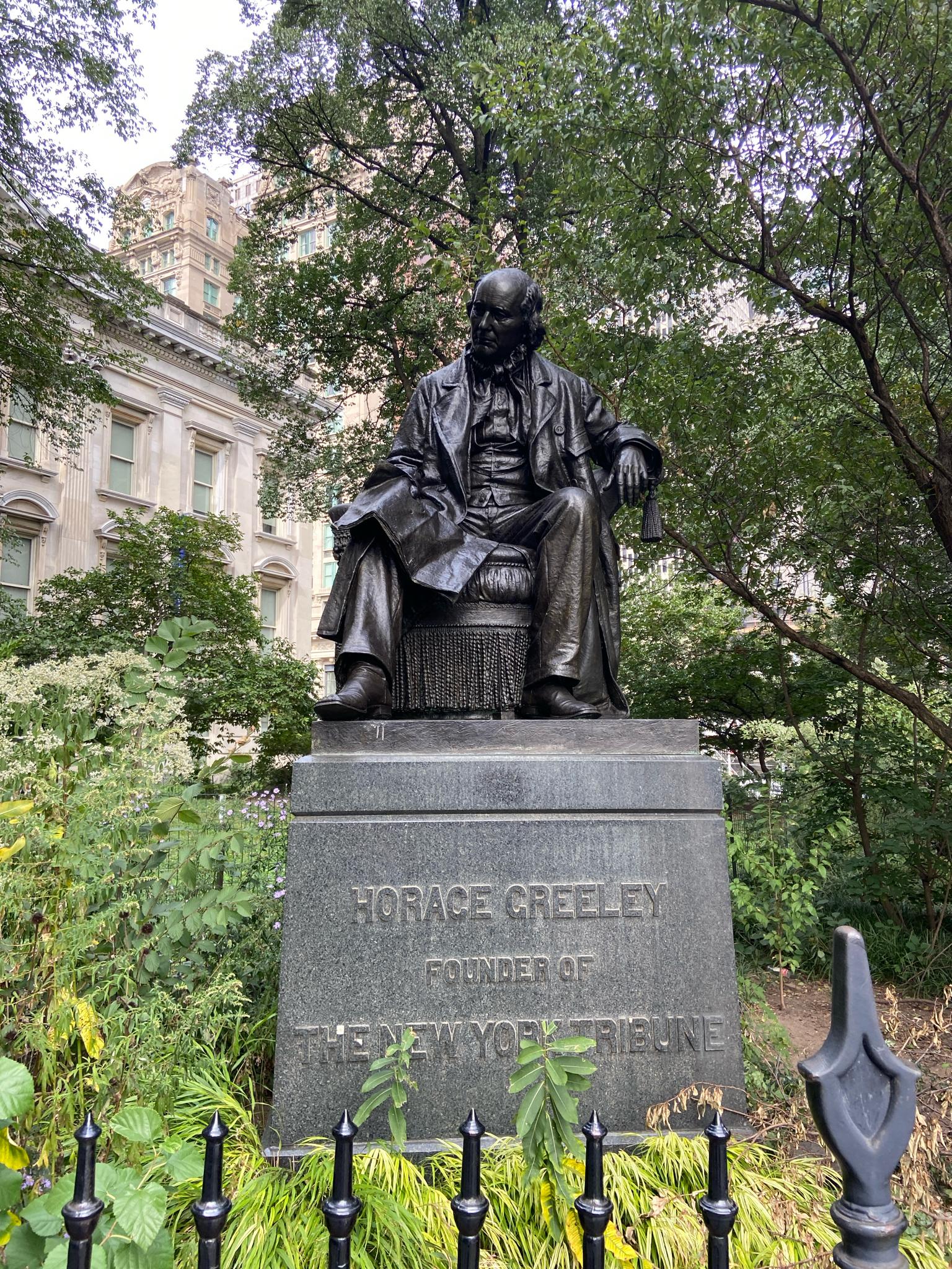 Bronze statue of a seated man on a stone pedestal, surrounded by greenery.
