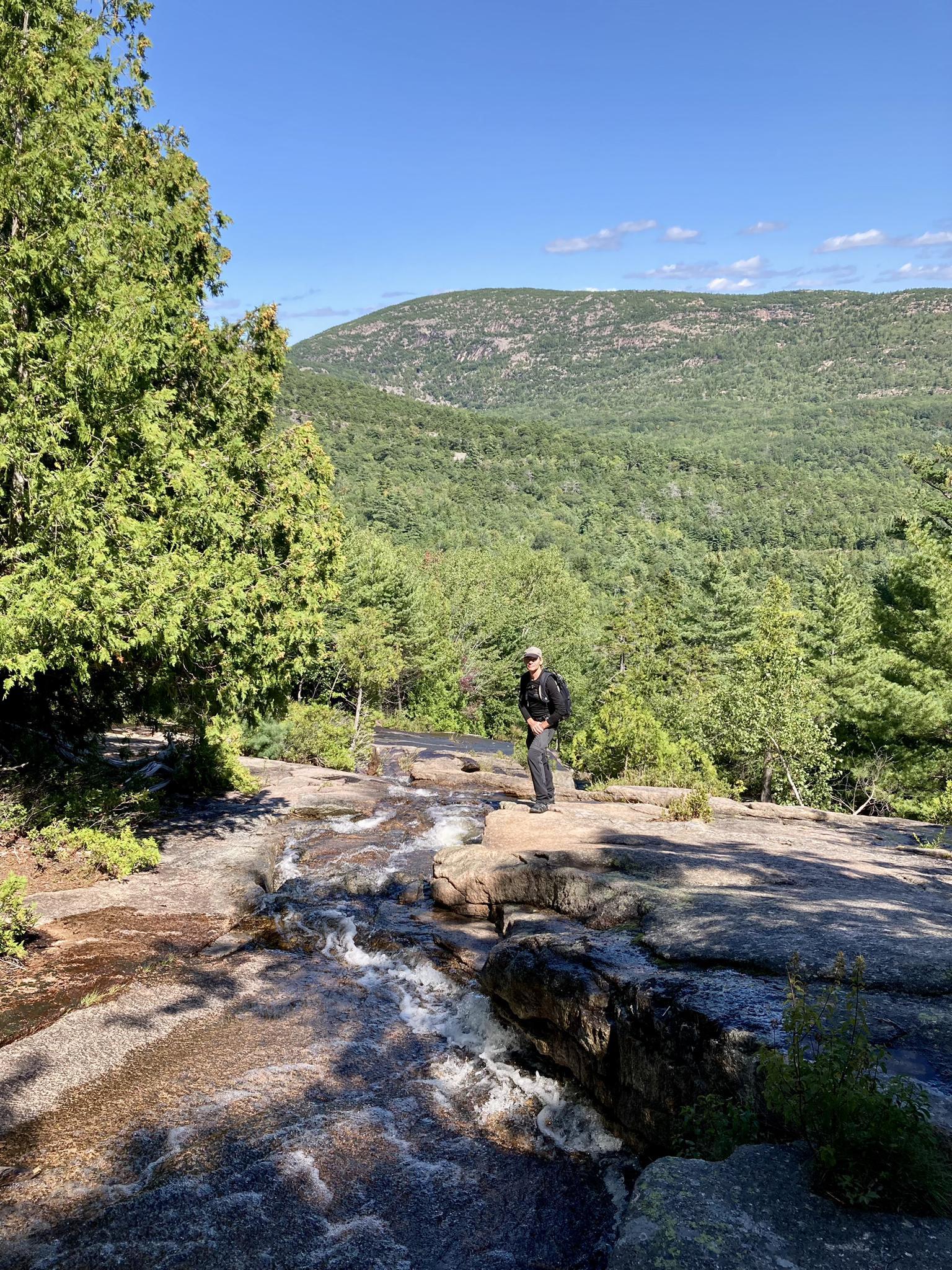 Person standing beside a small waterfall cascading over rocks, sunny day.