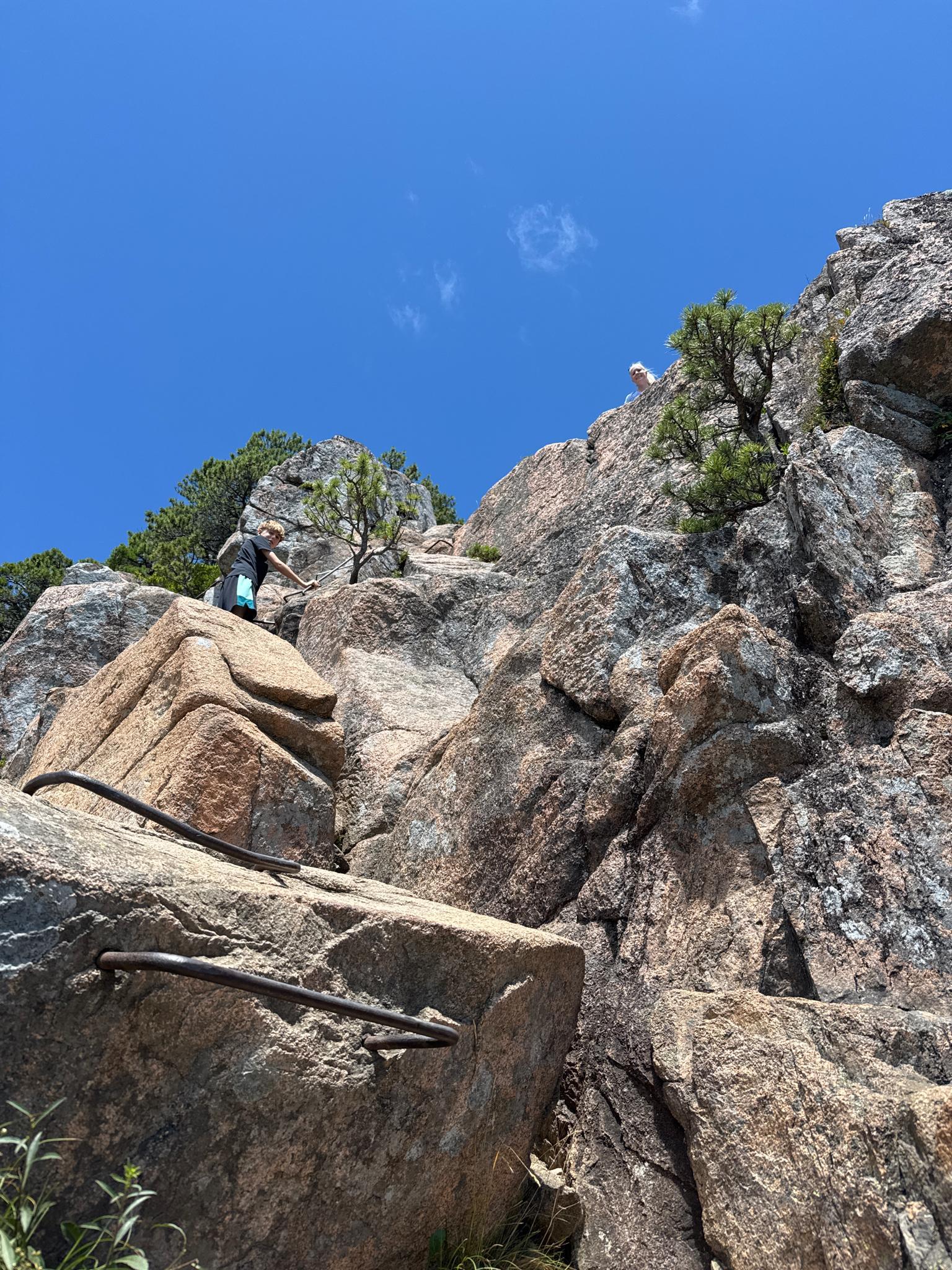 Two people climbing a steep rocky path with metal rungs under bright sun.