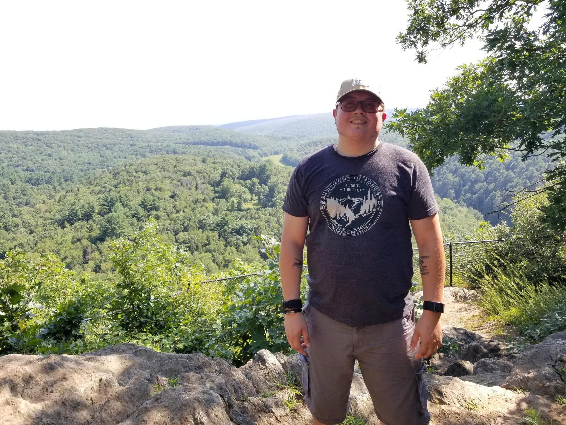 A man smiles at a scenic overlook with a vast green forest below.