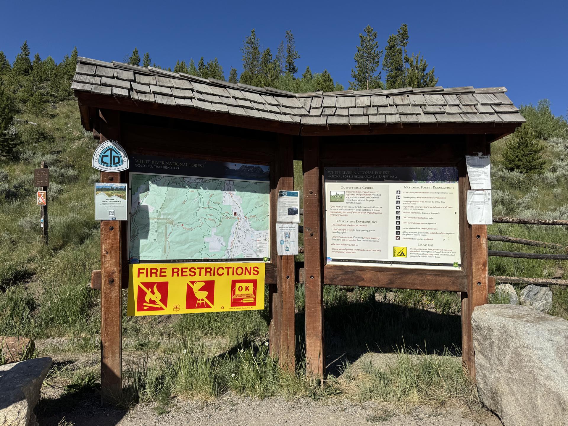 Wooden information kiosk with map and fire restrictions sign on a sunny hillside.