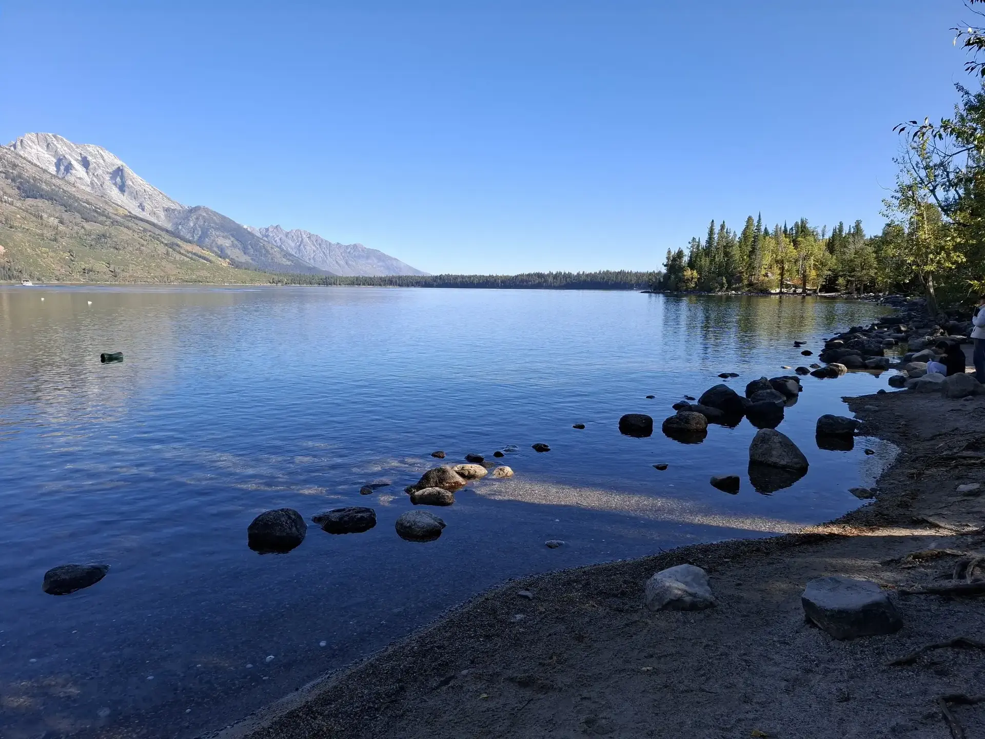 Clear lake with rocky shore, distant mountains, and blue sky.